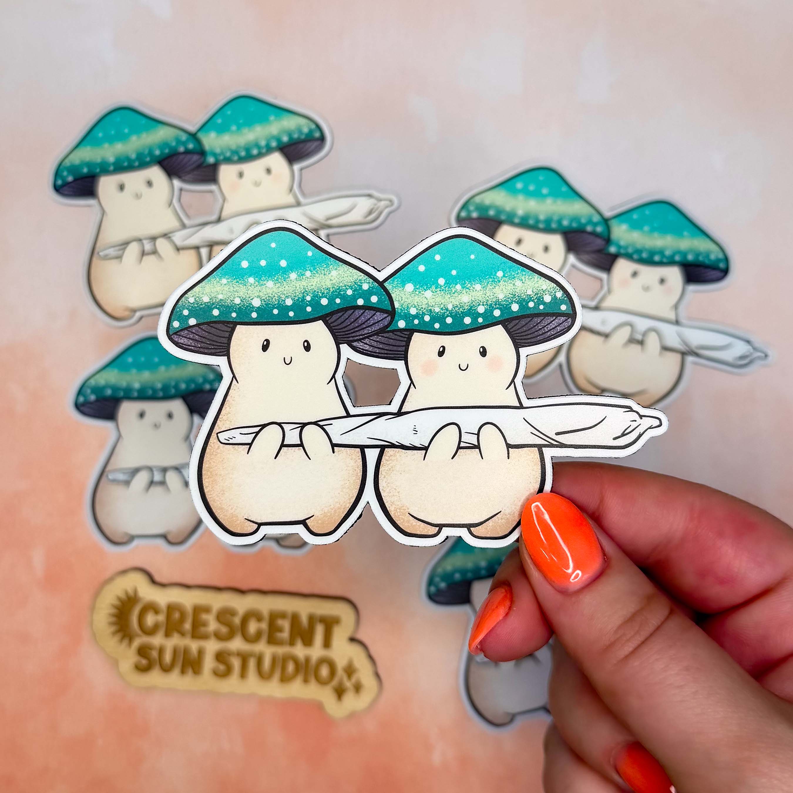 Two cream mushroom characters happily stand next to one another holding a giant joint that is larger than the two of them together! One mushroom has slightly rosier cheeks than the other. Both characters have a green mushroom cap and towards the bottom, the caps have a band of yellow and white dots that give a sparkle effect.

In this image, a hand holds a magnet of the joint venturers close to the camera while more Jay and Doobie magnets are slightly blurred in the background.
