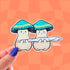Two cream mushroom characters happily stand next to one another holding a giant joint that is larger than the two of them together! One mushroom has slightly rosier cheeks than the other. Both characters have a green mushroom cap and towards the bottom, the caps have a band of yellow and white dots that give a sparkle effect.