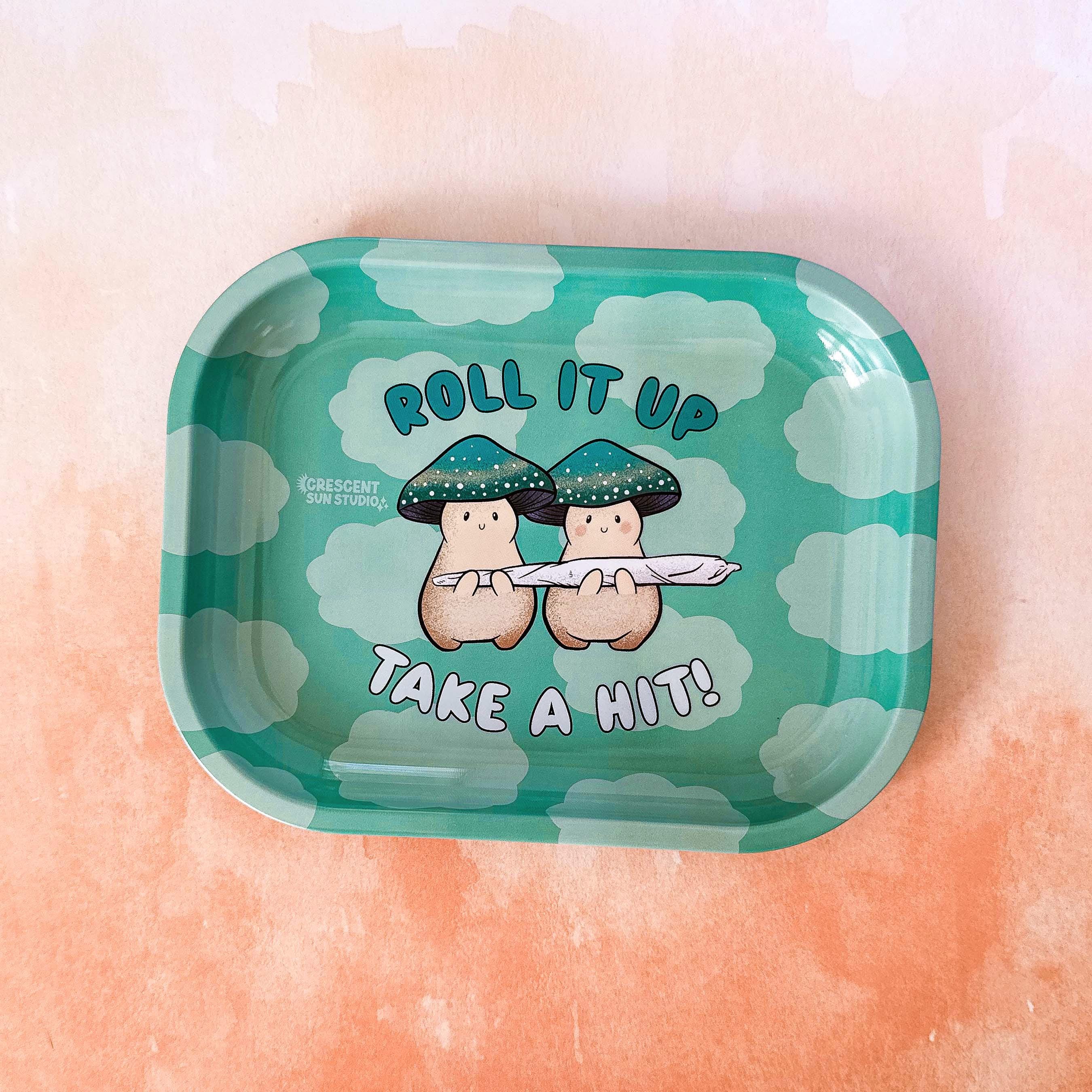 A small rolling tray with a teal cloud background. In the foreground,  text reads "roll it up, take a hit" and two cream mushroom characters happily stand next to one another holding a giant joint that is larger than the two of them together! One mushroom has slightly rosier cheeks than the other. Both characters have a green mushroom cap and towards the bottom, the caps have a band of yellow and white dots that give a sparkle effect. 