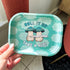 A small rolling tray with a teal cloud background. In the foreground,  text reads "roll it up, take a hit" and two cream mushroom characters happily stand next to one another holding a giant joint that is larger than the two of them together! One mushroom has slightly rosier cheeks than the other. Both characters have a green mushroom cap and towards the bottom, the caps have a band of yellow and white dots that give a sparkle effect. 