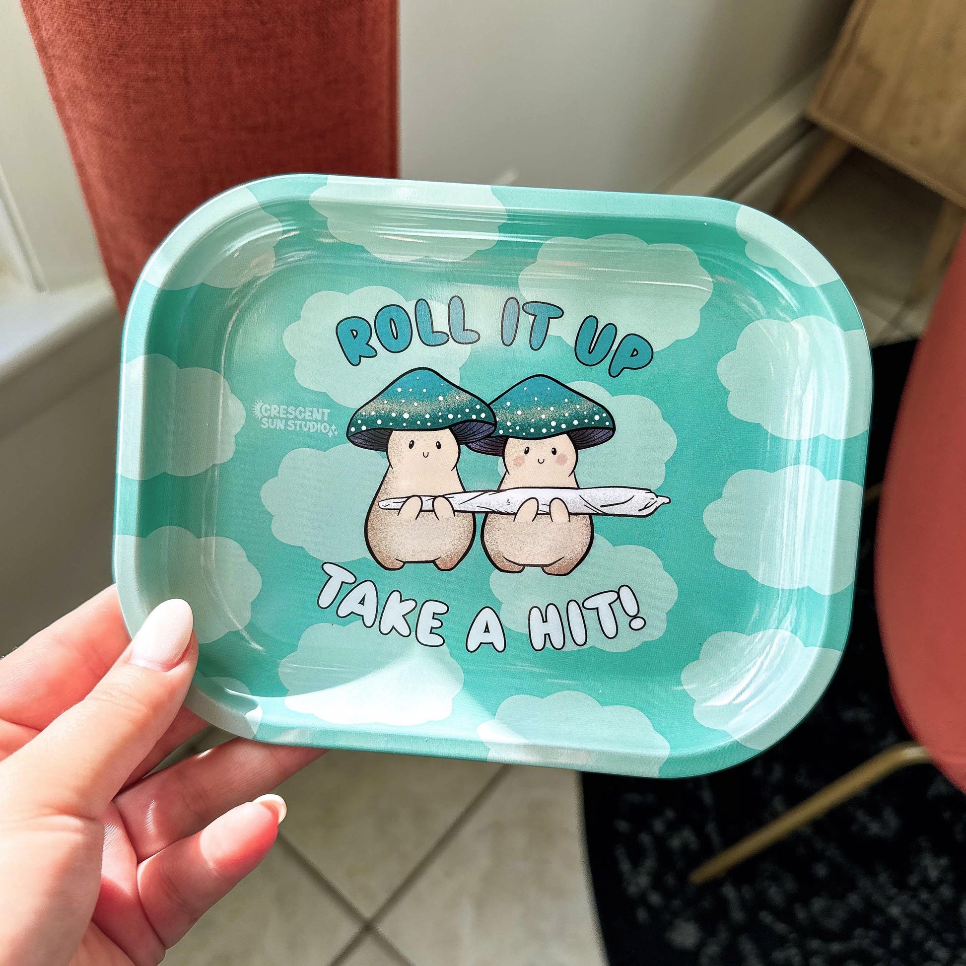 A small rolling tray with a teal cloud background. In the foreground,  text reads "roll it up, take a hit" and two cream mushroom characters happily stand next to one another holding a giant joint that is larger than the two of them together! One mushroom has slightly rosier cheeks than the other. Both characters have a green mushroom cap and towards the bottom, the caps have a band of yellow and white dots that give a sparkle effect. 