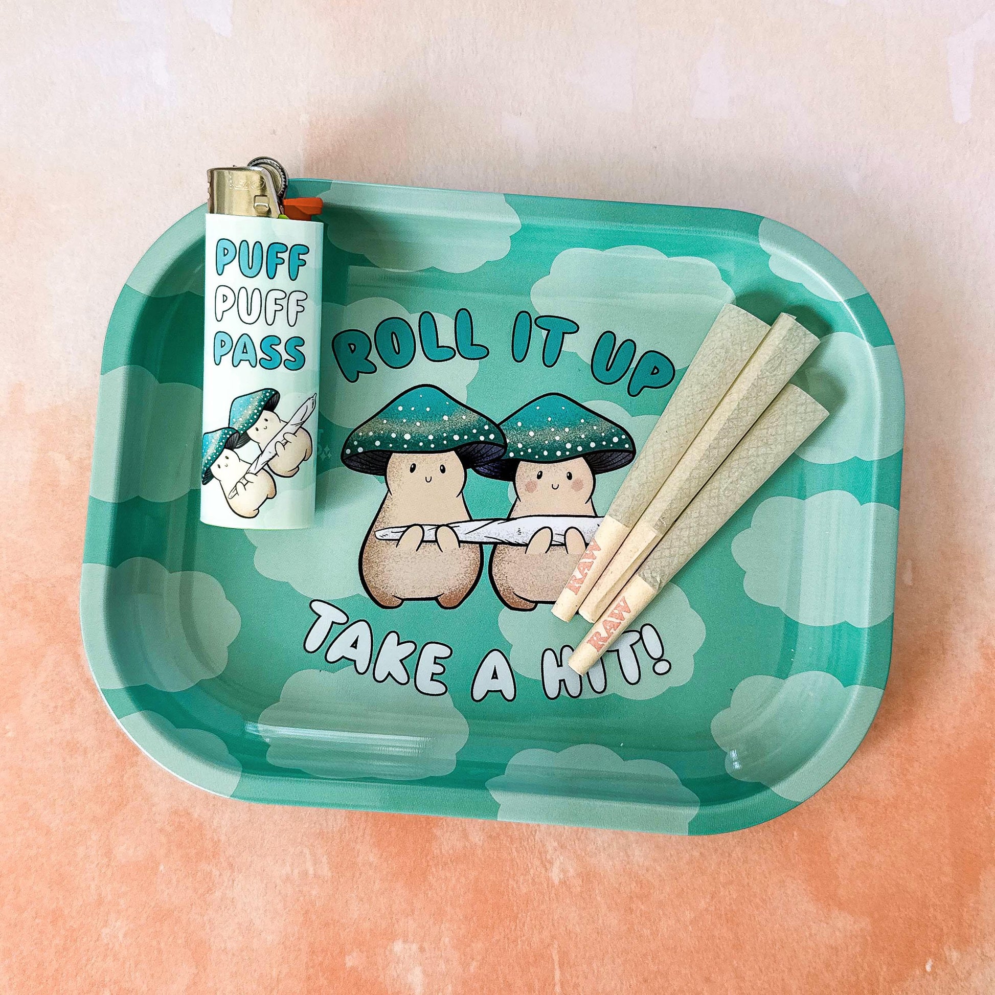 A small rolling tray with a teal cloud background. In the foreground,  text reads "roll it up, take a hit" and two cream mushroom characters happily stand next to one another holding a giant joint that is larger than the two of them together! One mushroom has slightly rosier cheeks than the other. Both characters have a green mushroom cap and towards the bottom, the caps have a band of yellow and white dots that give a sparkle effect. 

In this image, a lighter and pre-rolls sit on the tray