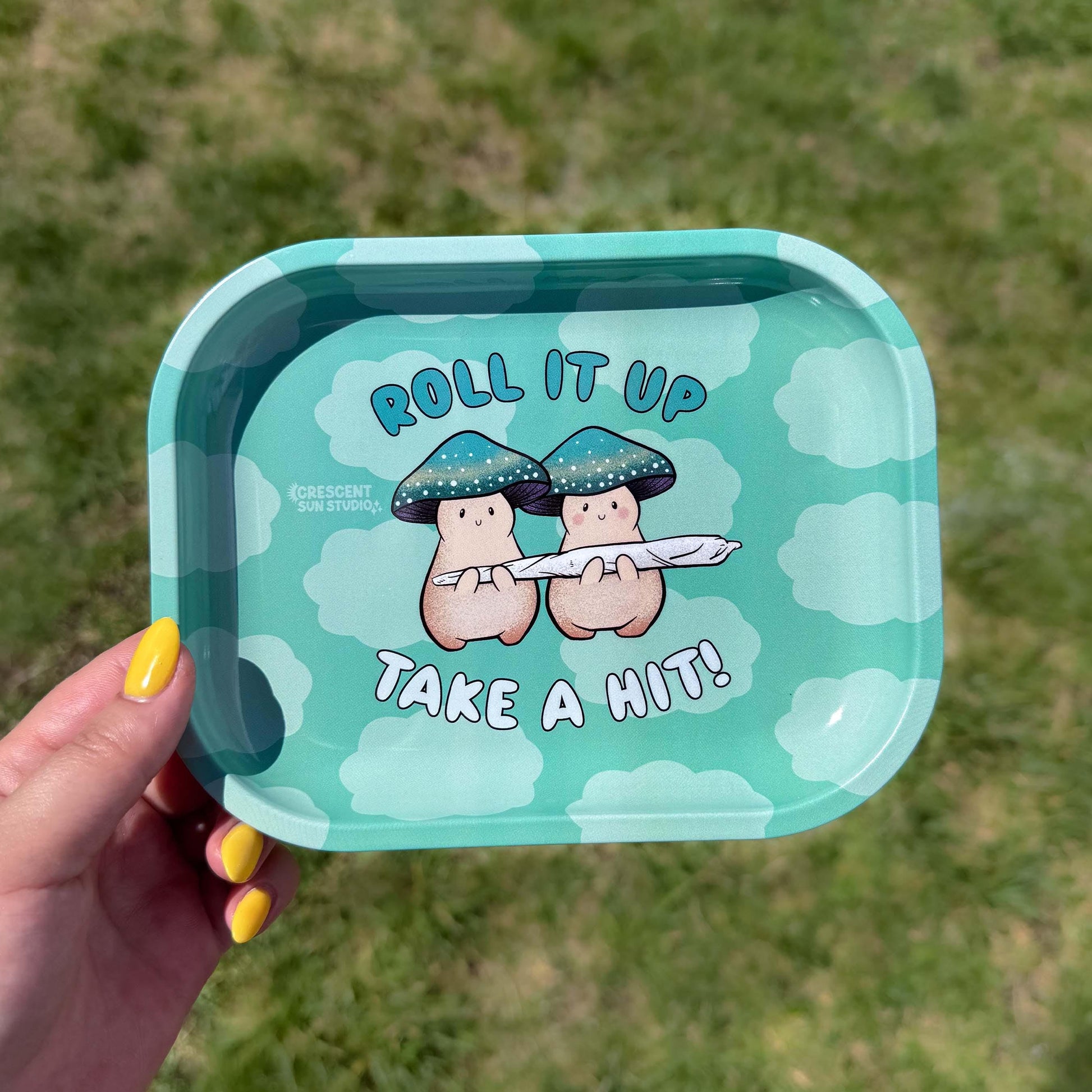 A small rolling tray with a teal cloud background. In the foreground,  text reads "roll it up, take a hit" and two cream mushroom characters happily stand next to one another holding a giant joint that is larger than the two of them together! One mushroom has slightly rosier cheeks than the other. Both characters have a green mushroom cap and towards the bottom, the caps have a band of yellow and white dots that give a sparkle effect. 

in this image, the tray is held in a person's hand, showing the size
