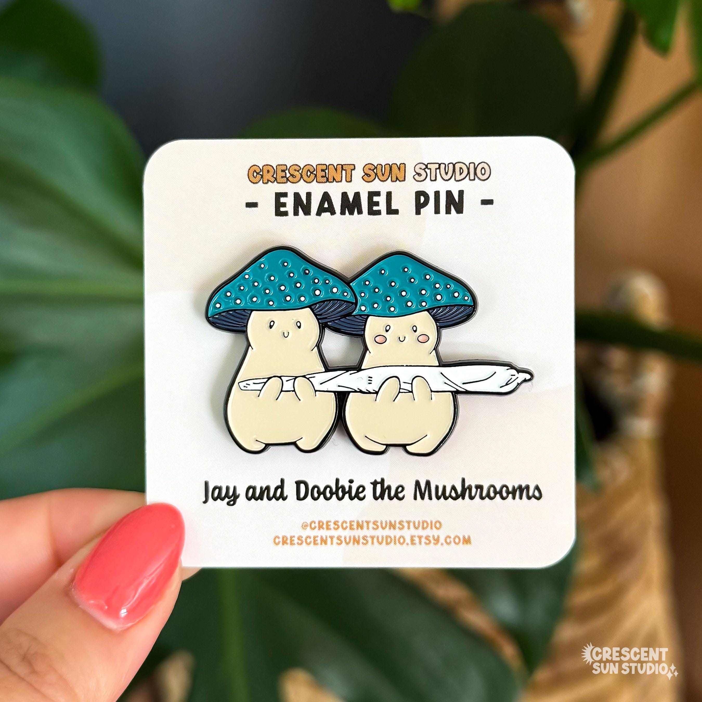 Two cream mushroom characters happily stand next to one another holding a giant joint that is larger than the two of them together! One mushroom has slightly rosier cheeks than the other. Both characters have a green mushroom cap and towards the bottom, the caps have a band of yellow and white dots that give a sparkle effect.

In this image, the pin version of the stoners is displayed on a light colored card and held close to the camera.