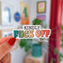 A die cut sticker with black text on the top that says "Kindly" and then rainbow text on the bottom that reads "fuck off"