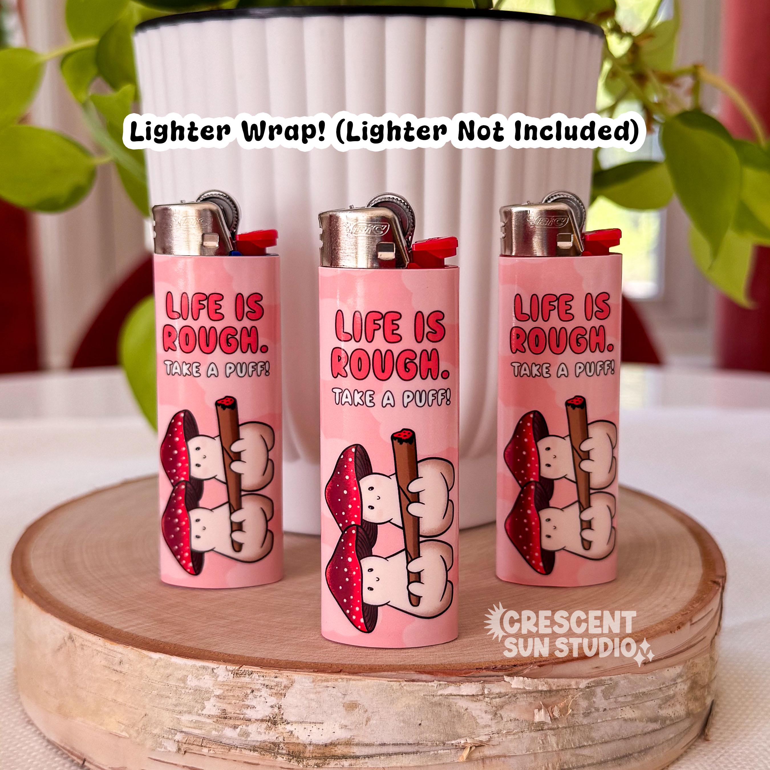 Three lighters are wrapped in a pink lighter wrap featuring two mushroom characters holding a joint. The mushrooms are pinky-beige and their caps are gradient-red with white spots. The text on each lighter reads "life is rough" written in a dark pink. Underneath in white text, it reads "take a puff." 

Across the image is the text, "lighter wrap! Lighter not included"