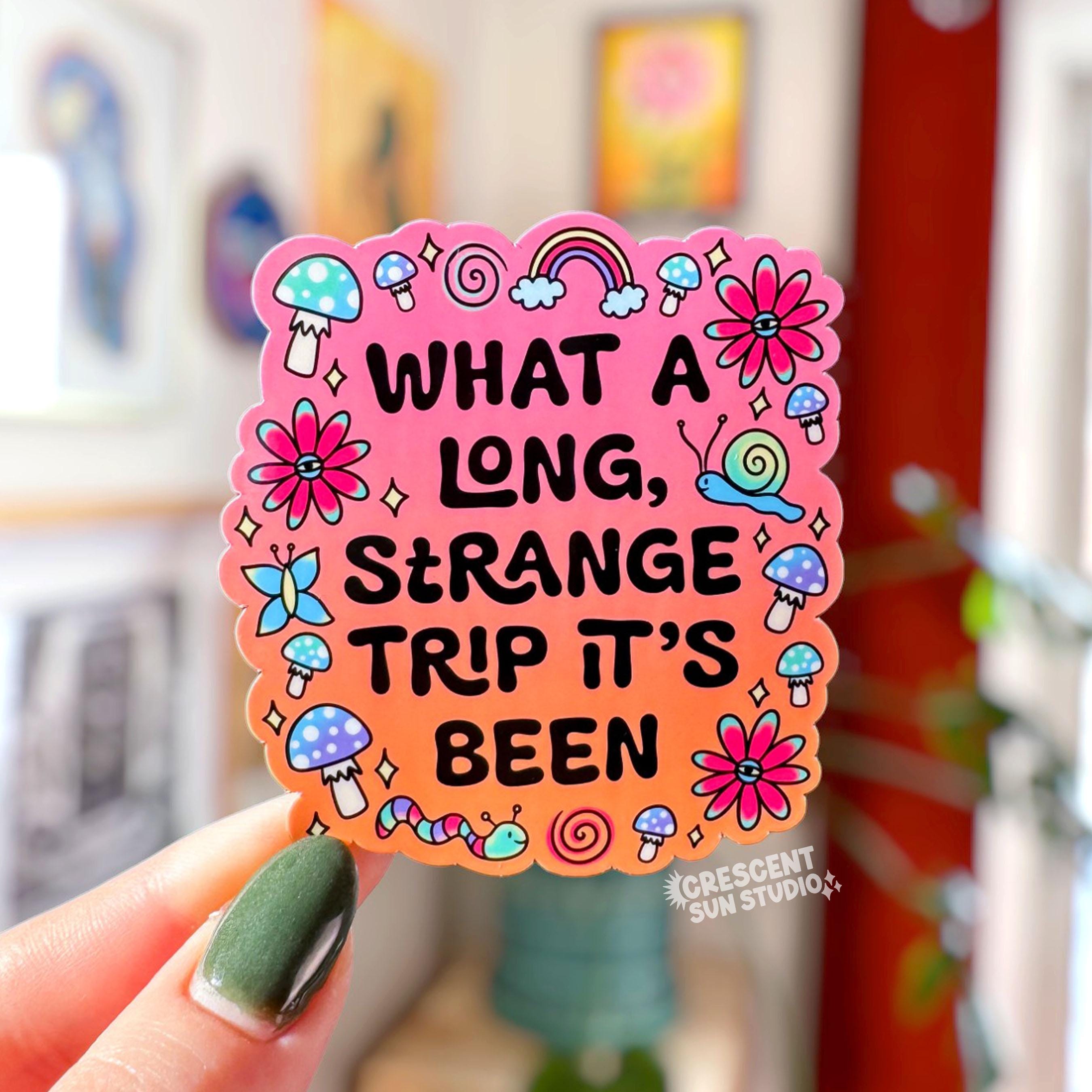 Die-cut sticker with a pink to orange gradient background reading 'What a Long Strange Trip It's Been' in black lettering surrounded by illustrated mushrooms, flowers with eyes, snails, a butterfly, and a worm.