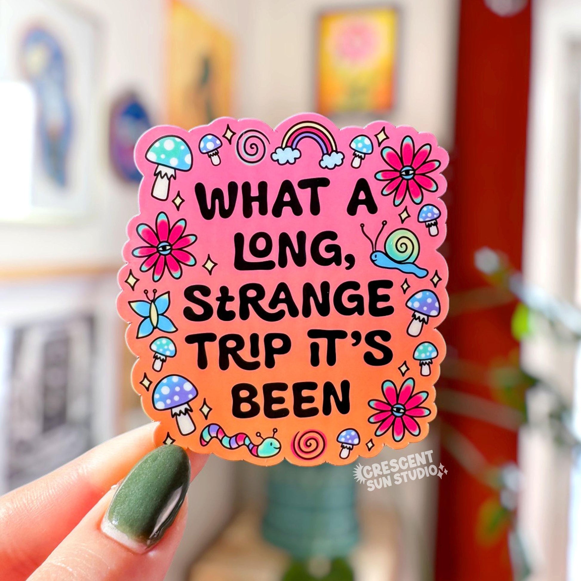 Die-cut sticker with a pink to orange gradient background reading 'What a Long Strange Trip It's Been' in black lettering surrounded by illustrated mushrooms, flowers with eyes, snails, a butterfly, and a worm.
