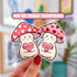 A hand holds two mushroom shaped die cut stickers. One is a happy and plump cream mushroom character with a pink cap covered in white hearts, holding a pink candy heart that reads "Mush Love 4 U." Lovey has some peachy-beige shading around their feet and bottom.

The other is similar, but with a light colored pink cap with dark hearts. Their candy heart reads "Love U So Mush!"