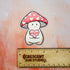 A magnet of Lovey, a happy and plump cream mushroom character with a pink cap covered in white hearts, holding a pink candy heart that reads "Mush Love 4 U." Lovey has some peachy-beige shading around their feet and bottom.

This photo shows Lovey above a wooden ruler, showing their size of 2.3 x 2.8 inches.