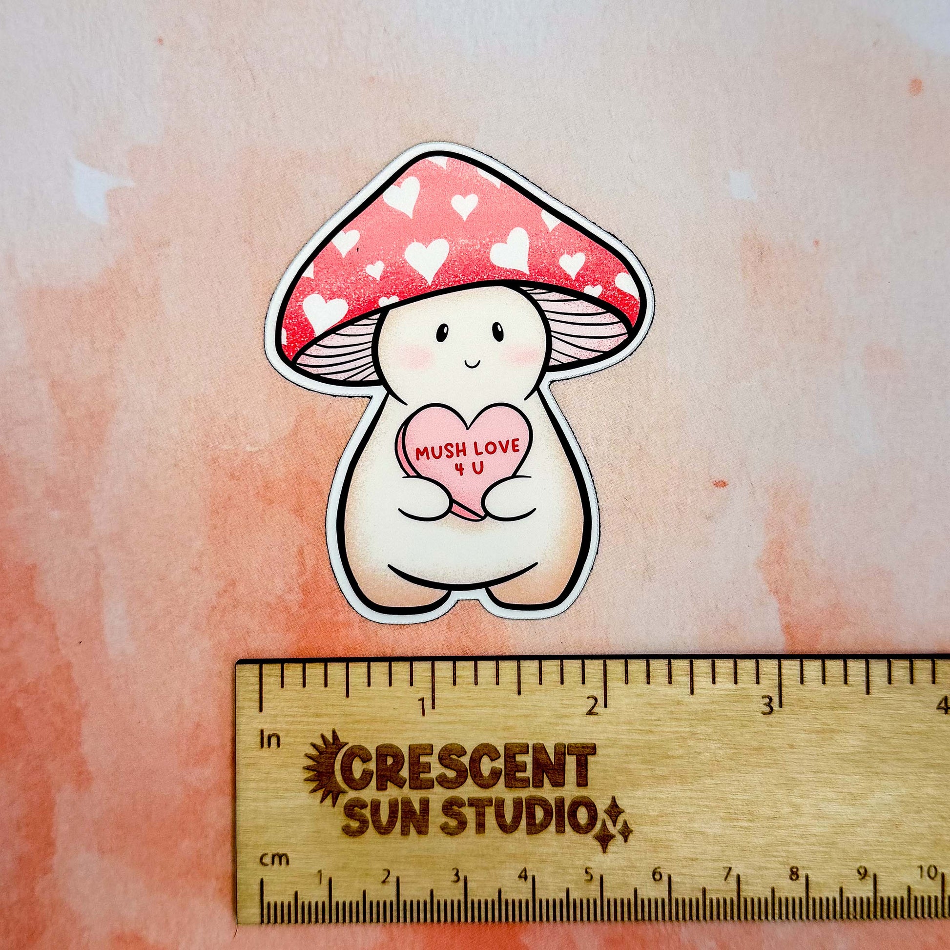 A magnet of Lovey, a happy and plump cream mushroom character with a pink cap covered in white hearts, holding a pink candy heart that reads "Mush Love 4 U." Lovey has some peachy-beige shading around their feet and bottom.

This photo shows Lovey above a wooden ruler, showing their size of 2.3 x 2.8 inches.