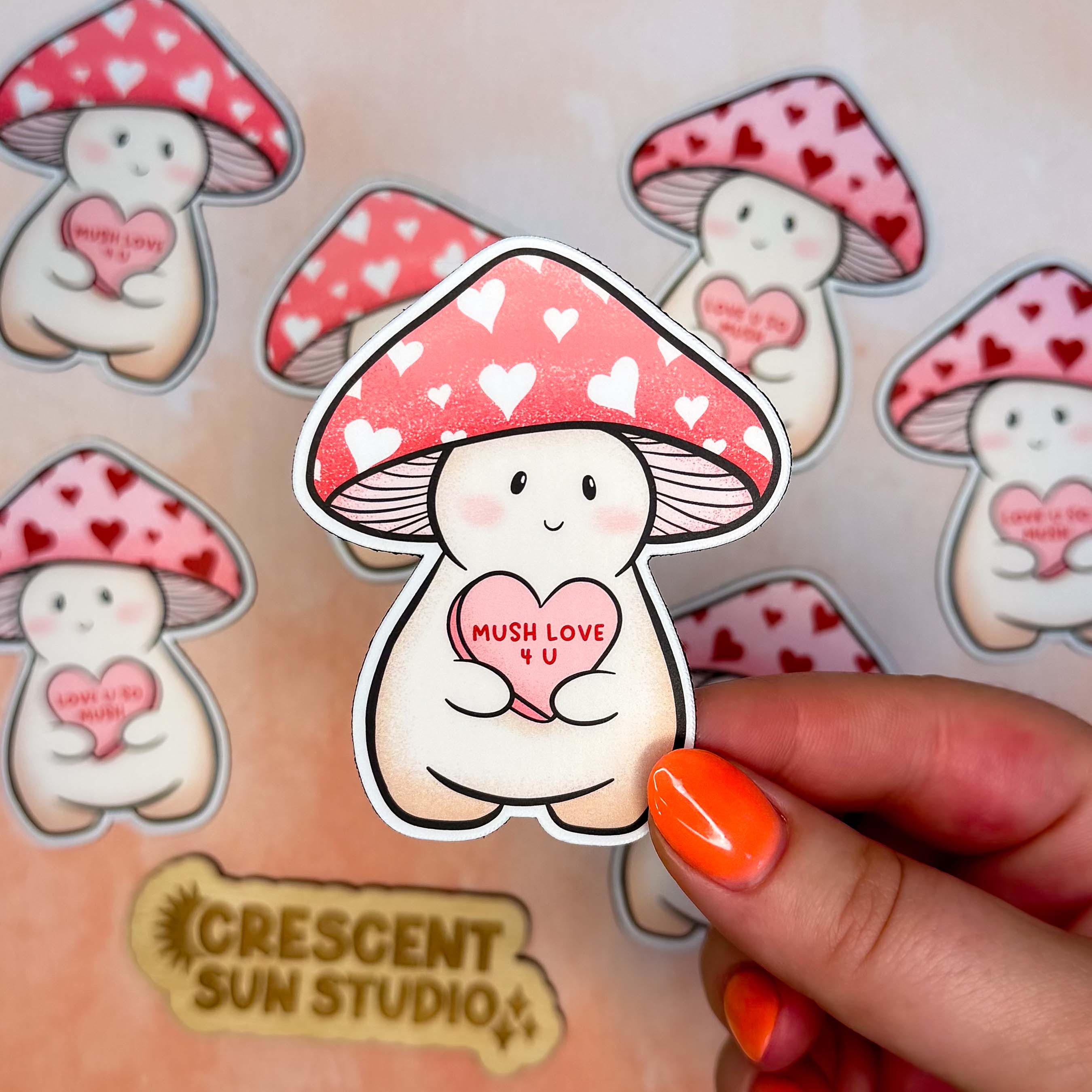 A hand holds one Lovey magnet close to the camera while there are other Lovey magnets in the background. Lovey is a happy and plump cream mushroom character with a pink cap covered in white hearts, holding a pink candy heart that reads "Mush Love 4 U." Lovey has some peachy-beige shading around their feet and bottom.