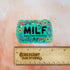 A bright green colored rectangle magnet with black bubble letters that says MILF and in smaller letters underneath that reads "man I love frolicking." The text is bordered by brightly colored mushrooms, rainbows, butterflies, and various flowers.

In this image, the magnet sits above a wooden ruler, showing its size of 3 inches.