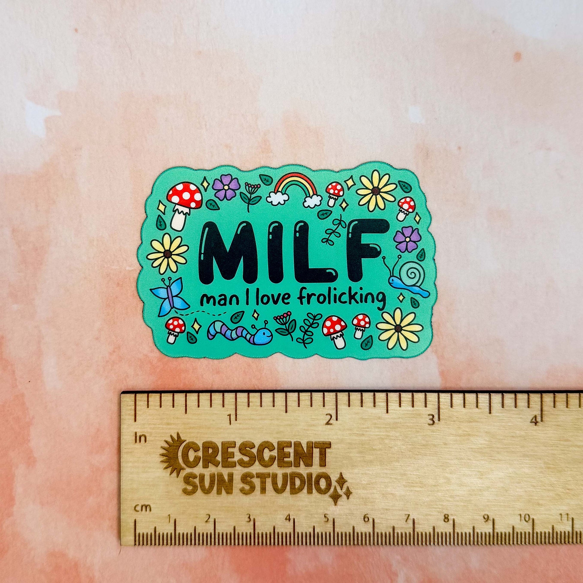 A bright green colored rectangle magnet with black bubble letters that says MILF and in smaller letters underneath that reads "man I love frolicking." The text is bordered by brightly colored mushrooms, rainbows, butterflies, and various flowers.

In this image, the magnet sits above a wooden ruler, showing its size of 3 inches.