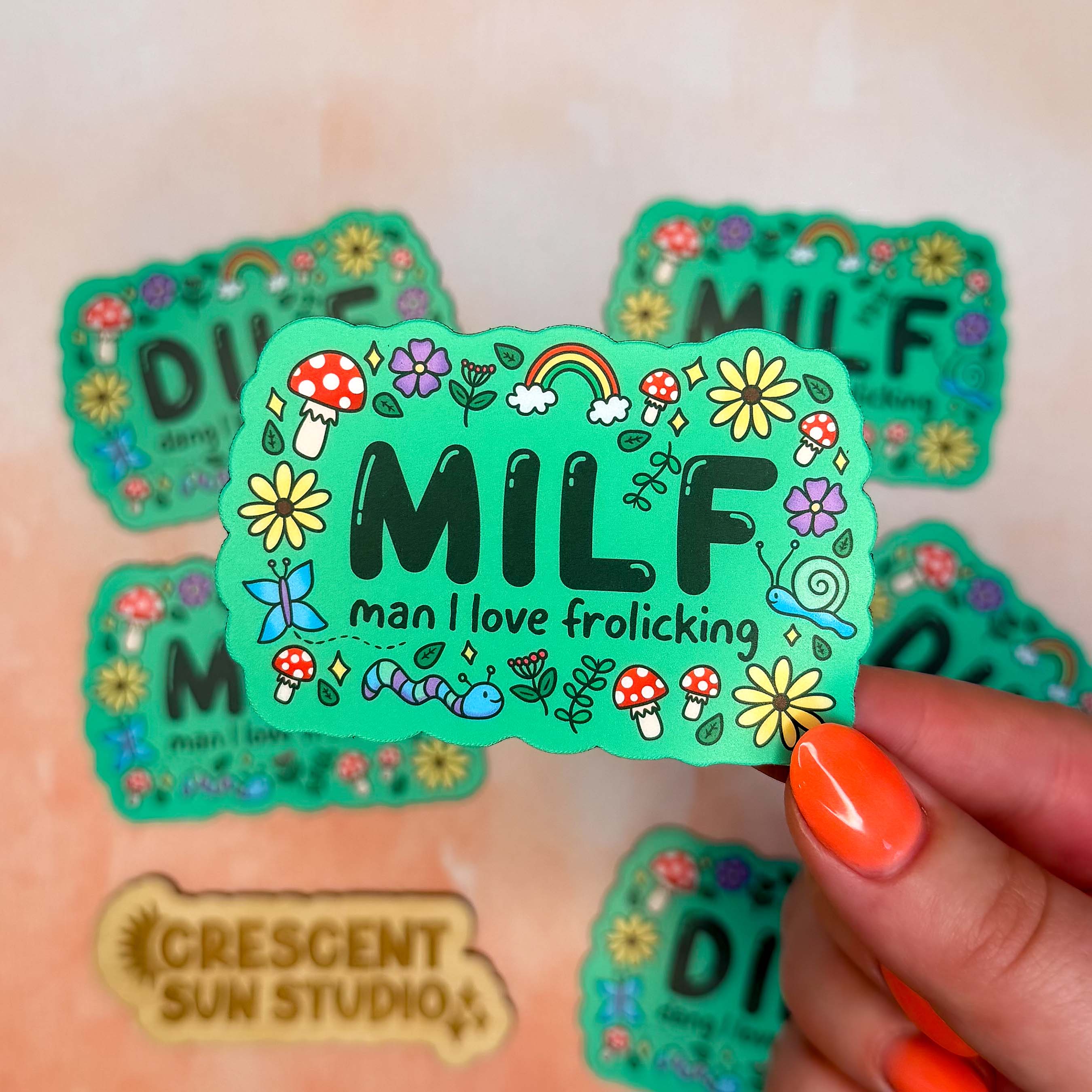 A bright green colored rectangle magnet with black bubble letters that says MILF and in smaller letters underneath that reads "man I love frolicking." The text is bordered by brightly colored mushrooms, rainbows, butterflies, and various flowers.

In this image, the magnet is held close to the camera with other magnets slightly blurred in the background.