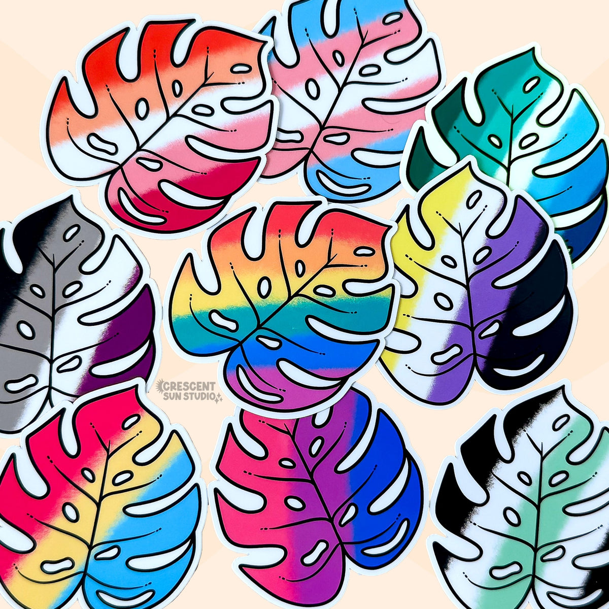 Colorful monstera leaf shaped stickers, each in the colors of different pride flags, are scattered on a beige surface.