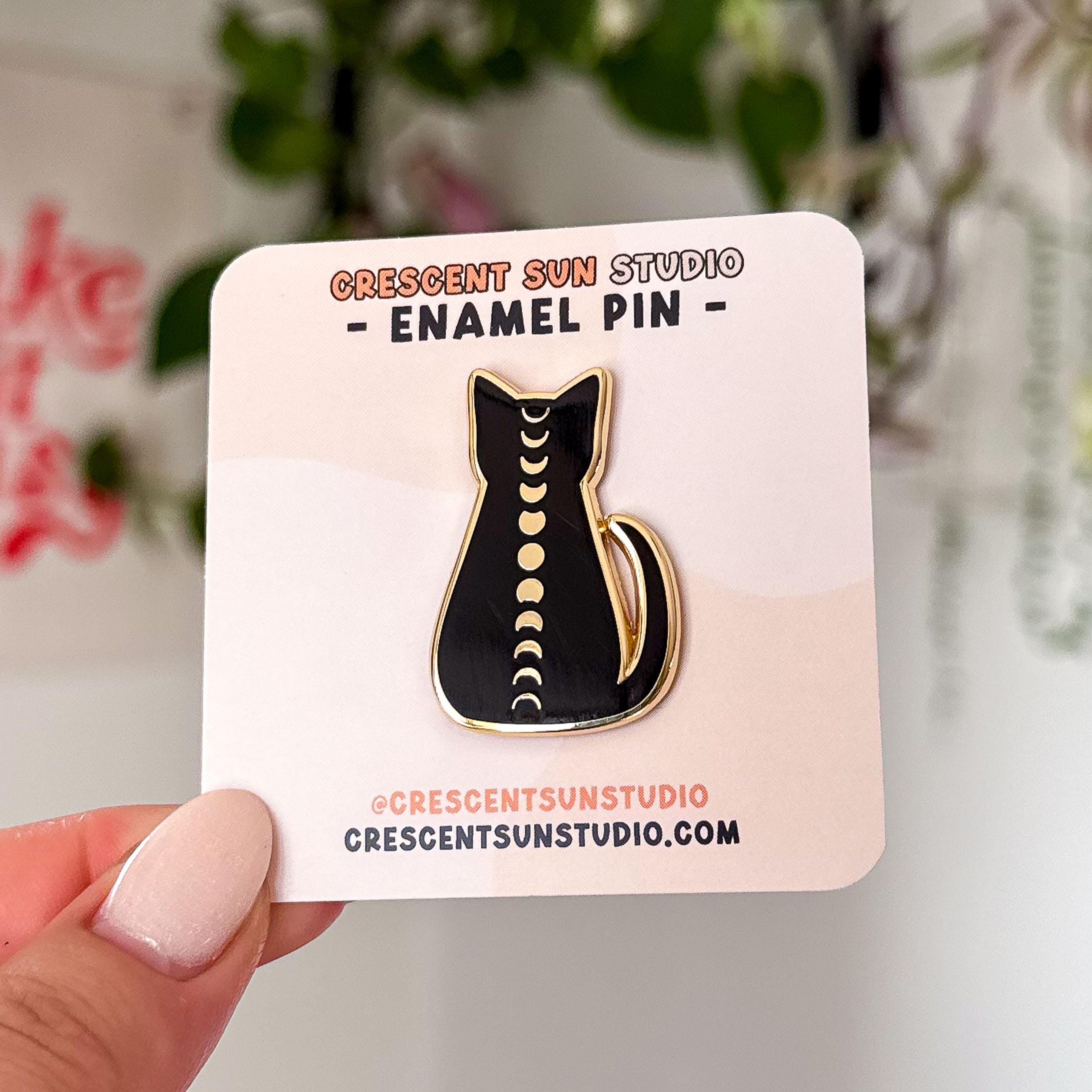This enamel pin is of a black cat silhouette trimmed in gold, with the full cycle of moon phases running down its spine.

In this image, the pin is displayed on a light colored card and held up to the camera.