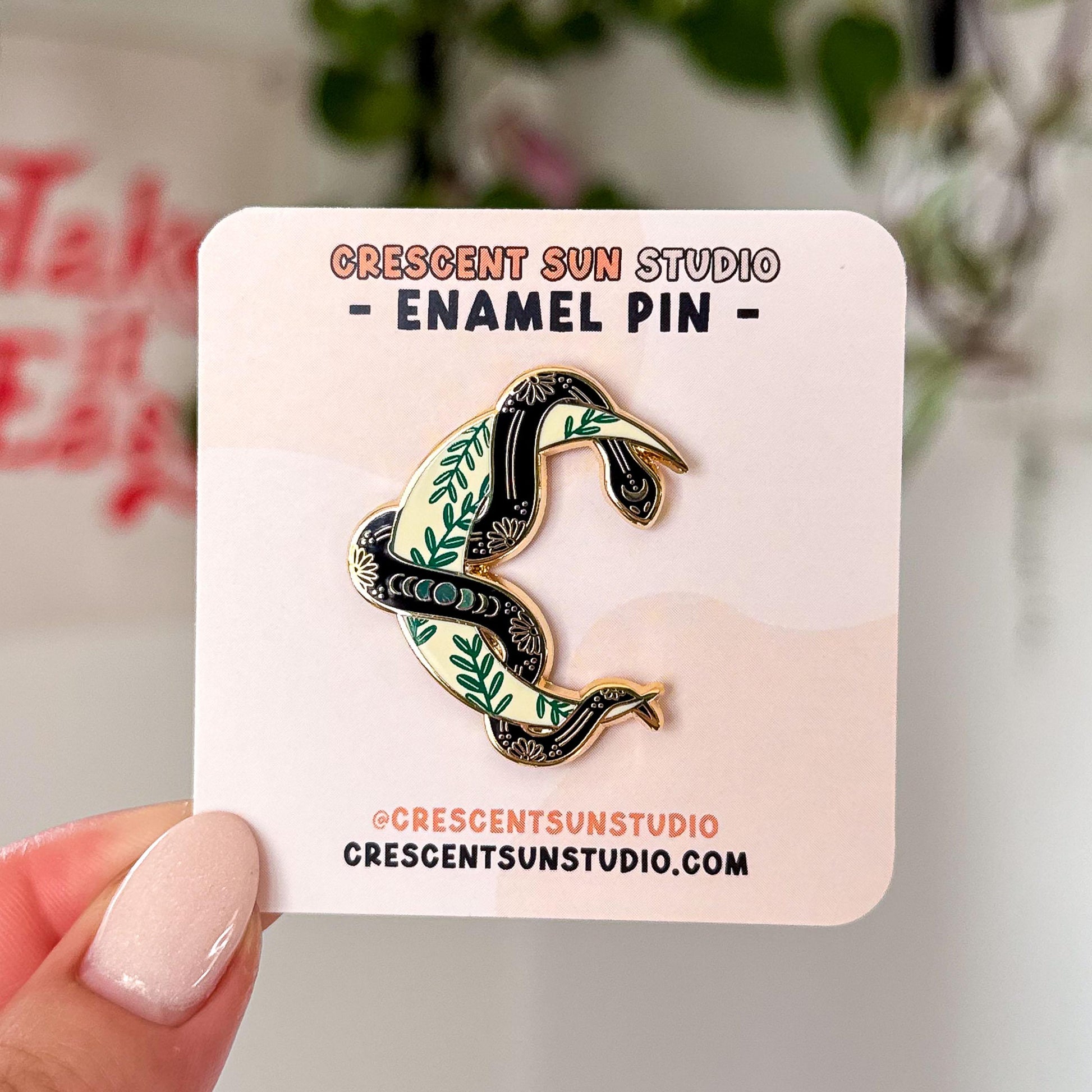An enamel pin, outlined in gold, in the shape of a yellow crescent moon with a black snake wrapped around it. The moon has a green ivy pattern, while the snake has several golden elements, like flowers and moon phases embeded into its skin.

This image shows the pin displayed on a light colored card and held close to the camera.