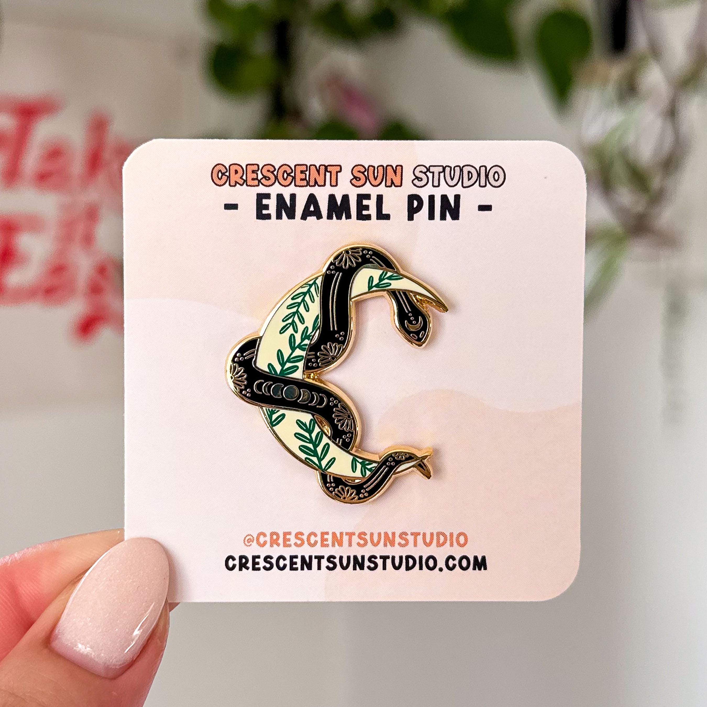 An enamel pin, outlined in gold, in the shape of a yellow crescent moon with a black snake wrapped around it. The moon has a green ivy pattern, while the snake has several golden elements, like flowers and moon phases embedded into its skin.

In this image, the pin is displayed on a light colored card and held up close to the camera.