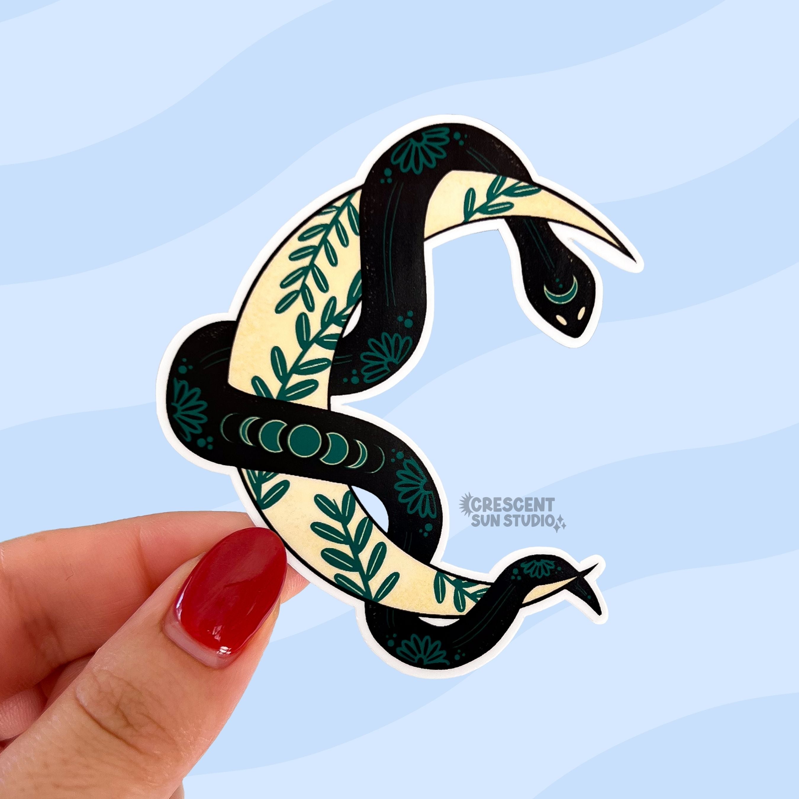 A sticker in the shape of a yellow crescent moon with a black snake wrapped around it is held close to the camera. The moon has a green ivy pattern, while the snake has light patterns in her skin, including moon phases toward the middle.