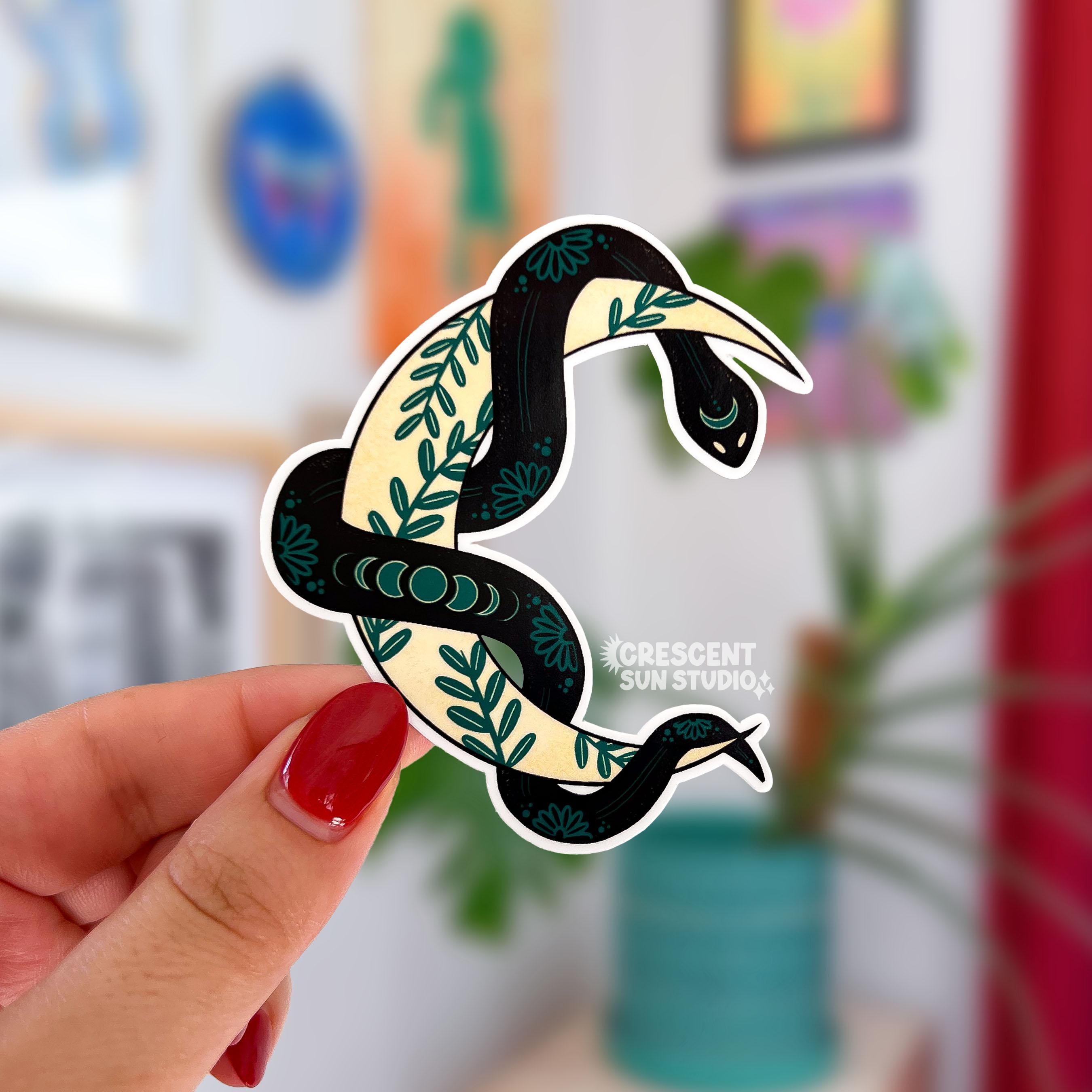 A sticker in the shape of a yellow crescent moon with a black snake wrapped around it is held close to the camera. The moon has a green ivy pattern, while the snake has light patterns in her skin, including moon phases toward the middle.
