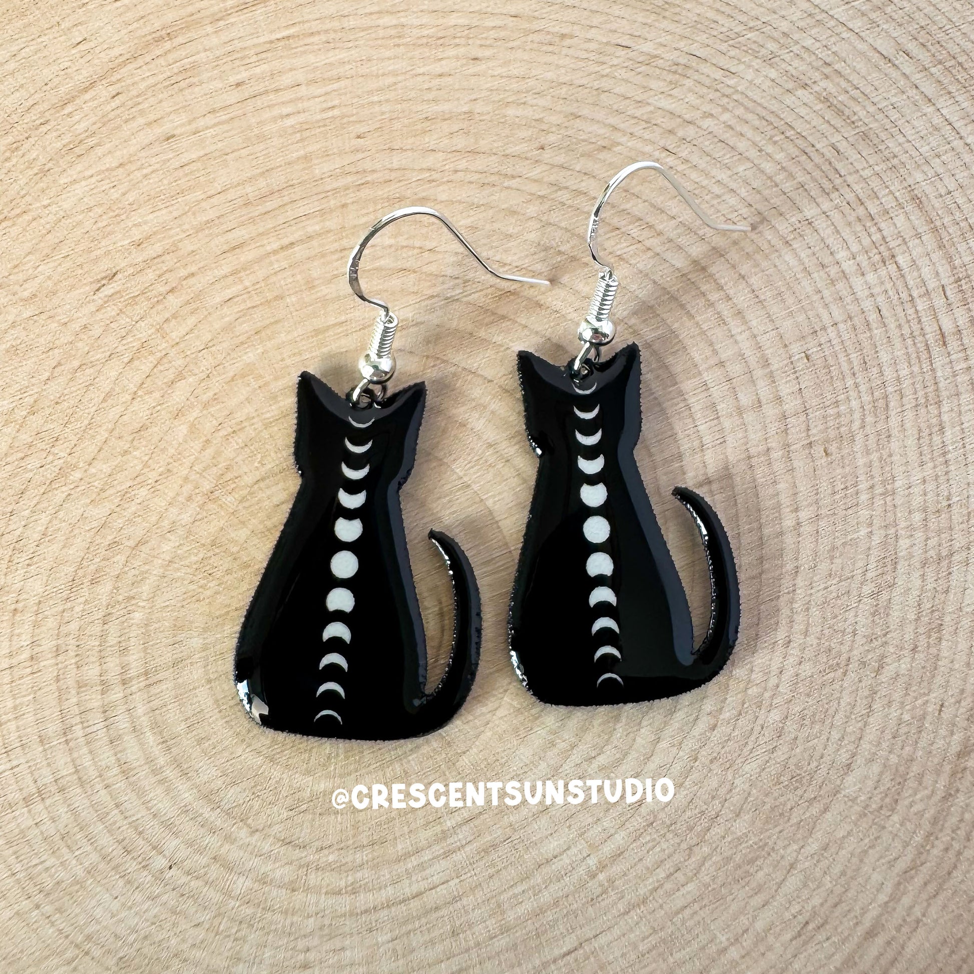 This image depicts a pair of dangle earrings. The hook for the earrings is silvertone. The earrings themselves depict a black cat silhouette lwith the full cycle of moon phases running down its spine.