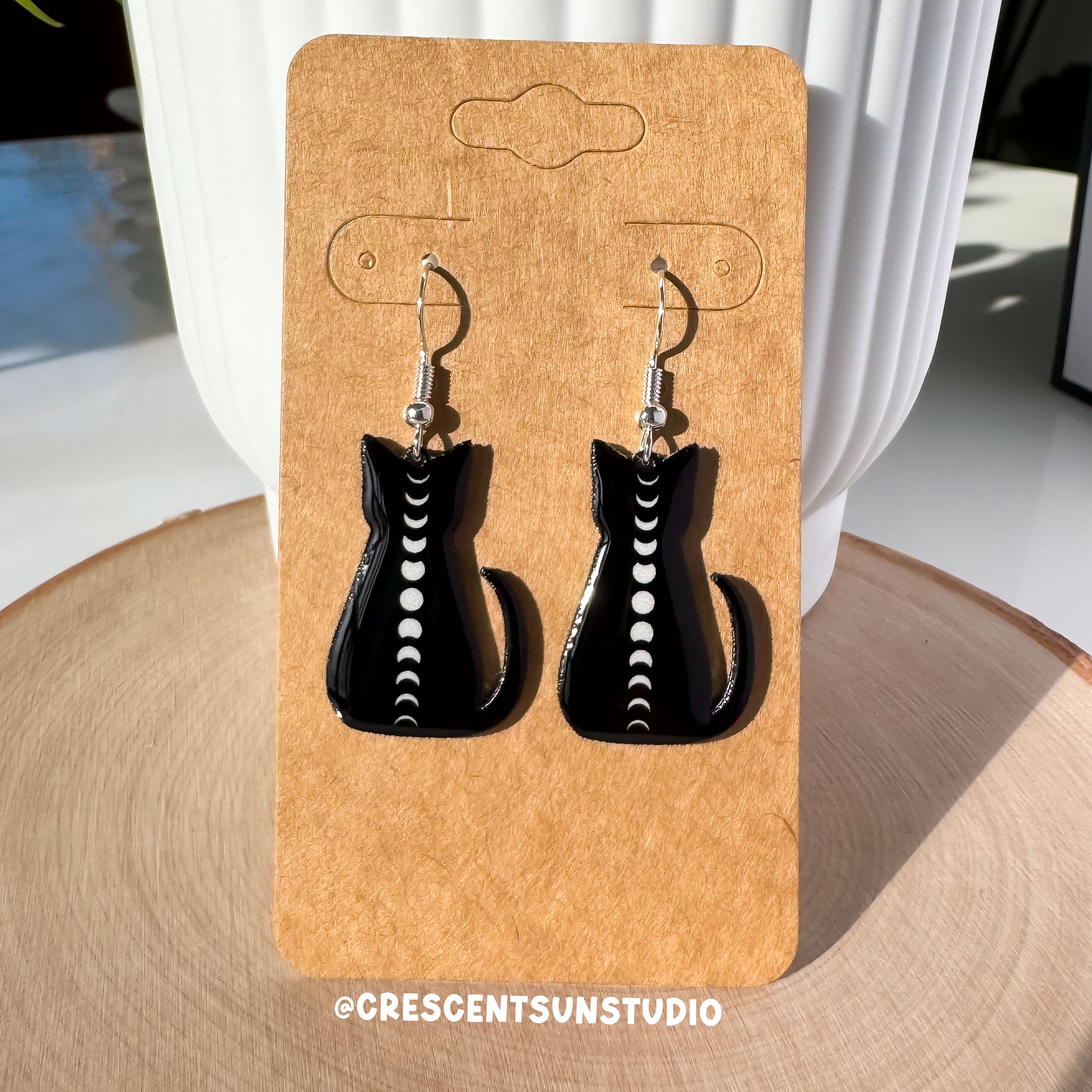 A pair of dangle earrings are displayed on a brown colored card. The hook for the earrings is silvertone. The earrings themselves depict a black cat silhouette lwith the full cycle of moon phases running down its spine.