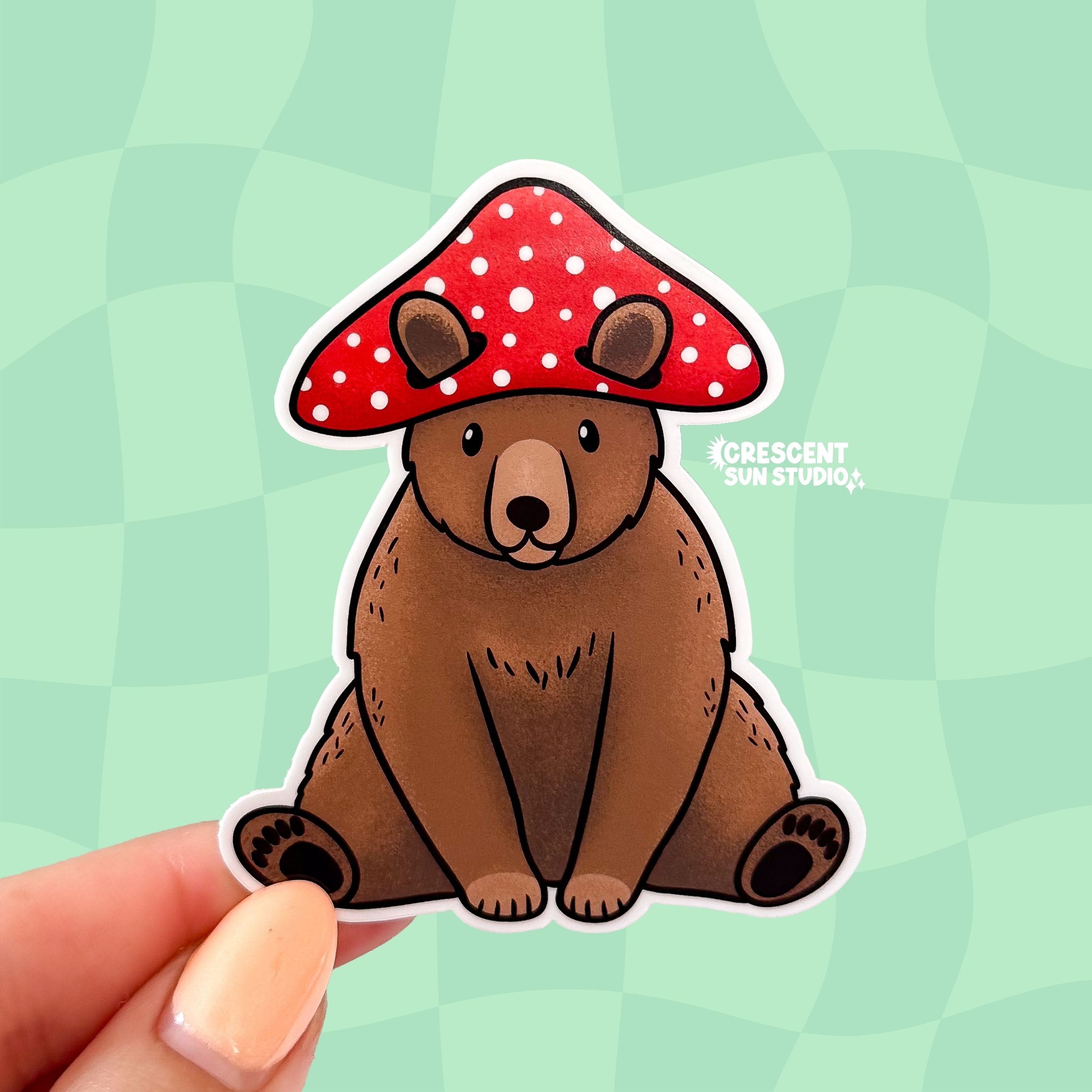 A die cut sticker of a brown bear sitting down and wearing a red and white mushroom hat.