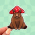 A die cut sticker of a brown bear sitting down and wearing a red and white mushroom hat.