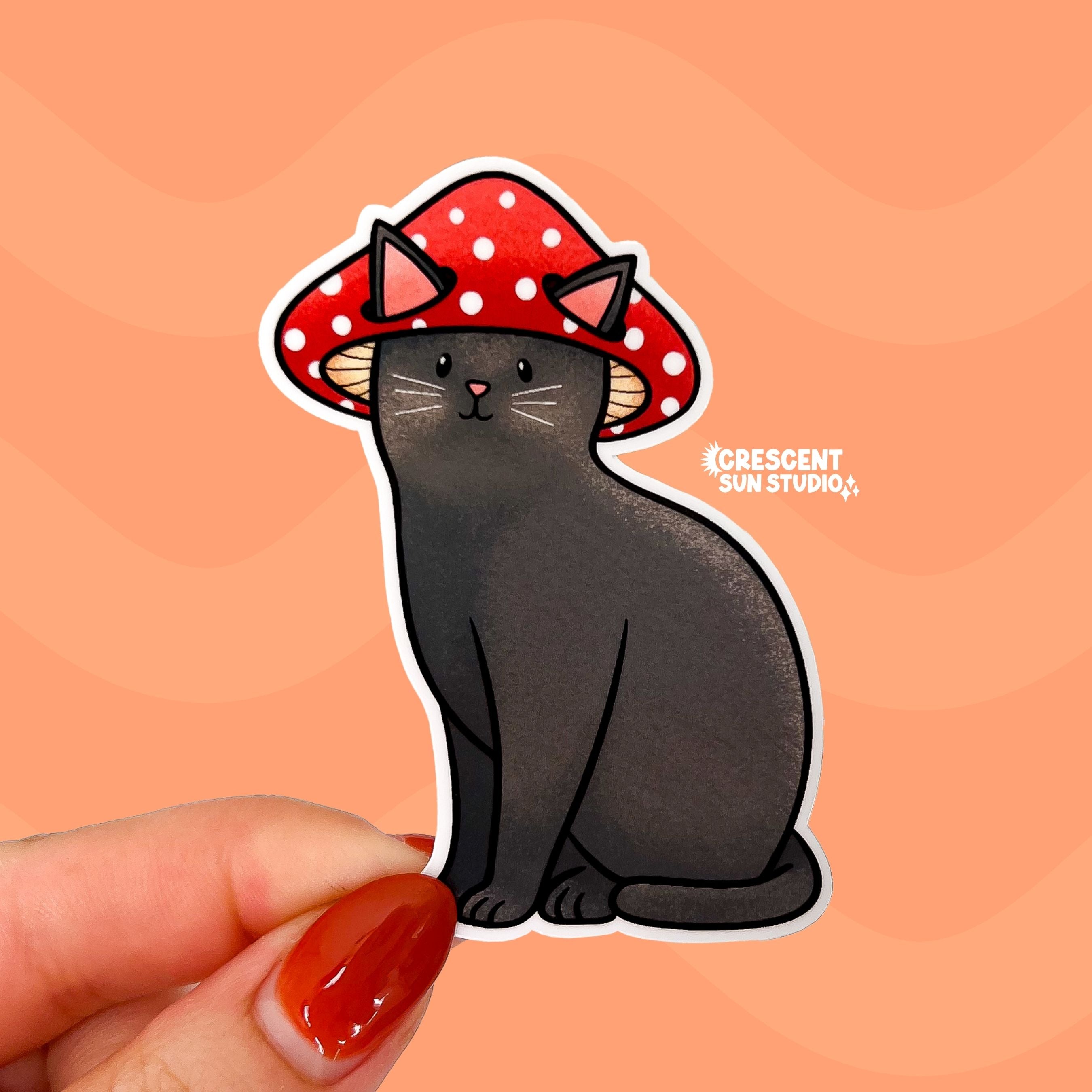 A sticker in the shape of a sitting cat is held close to the camera. The cat is black with white whiskers and has a contented look on his face. He is wearing a red mushroom cap with white spots and his large pink ears poke through holes in the cap.