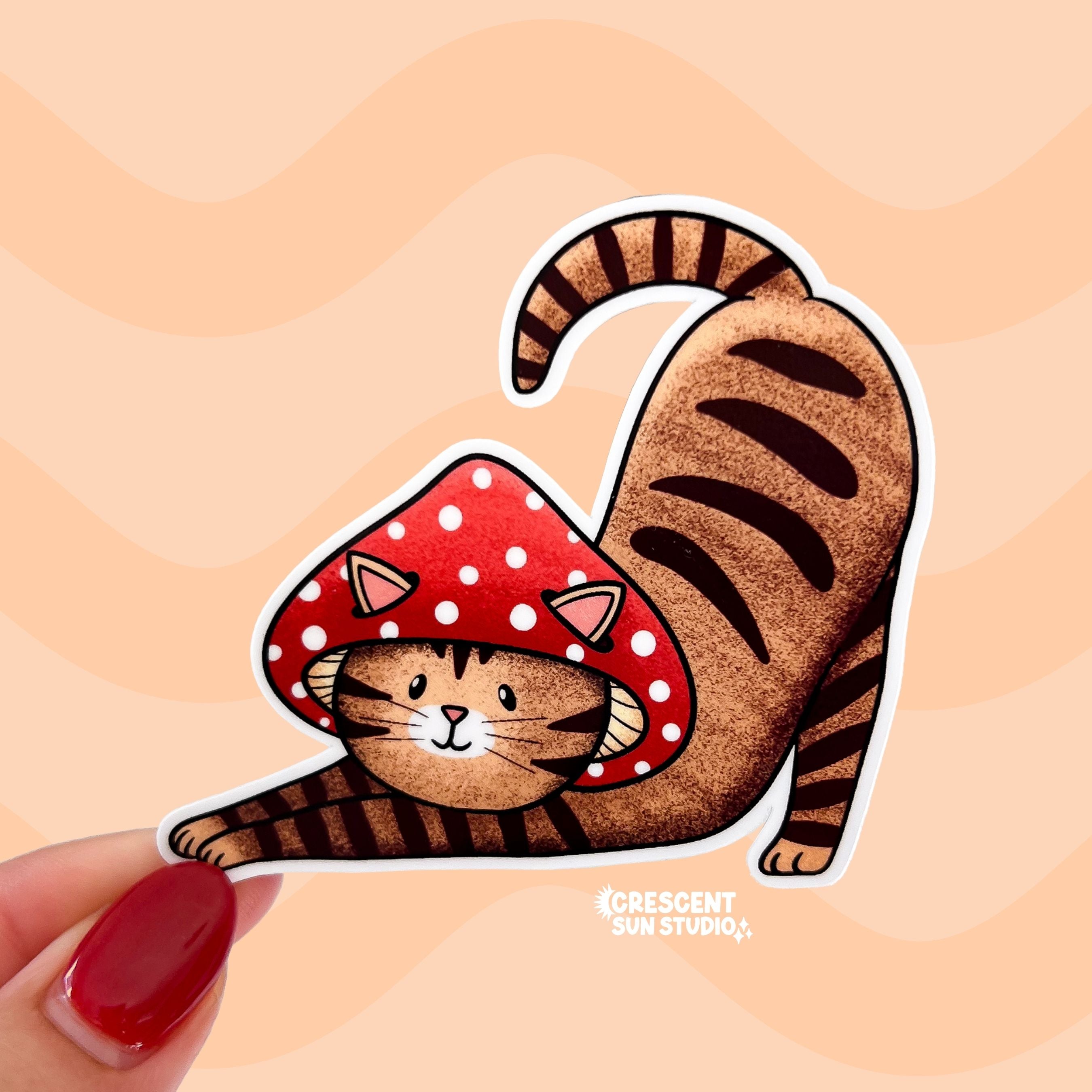 A sticker in the shape of a cat wearing a mushroom hat is held close to the camera. The cat is a brown tabby (medium brown with darker brown stripes) and has a sweet expression on his face. His red mushroom hat with white spots has spots for his ears to poke through. The cat is in a stretching pose with his tail curling up behind him.