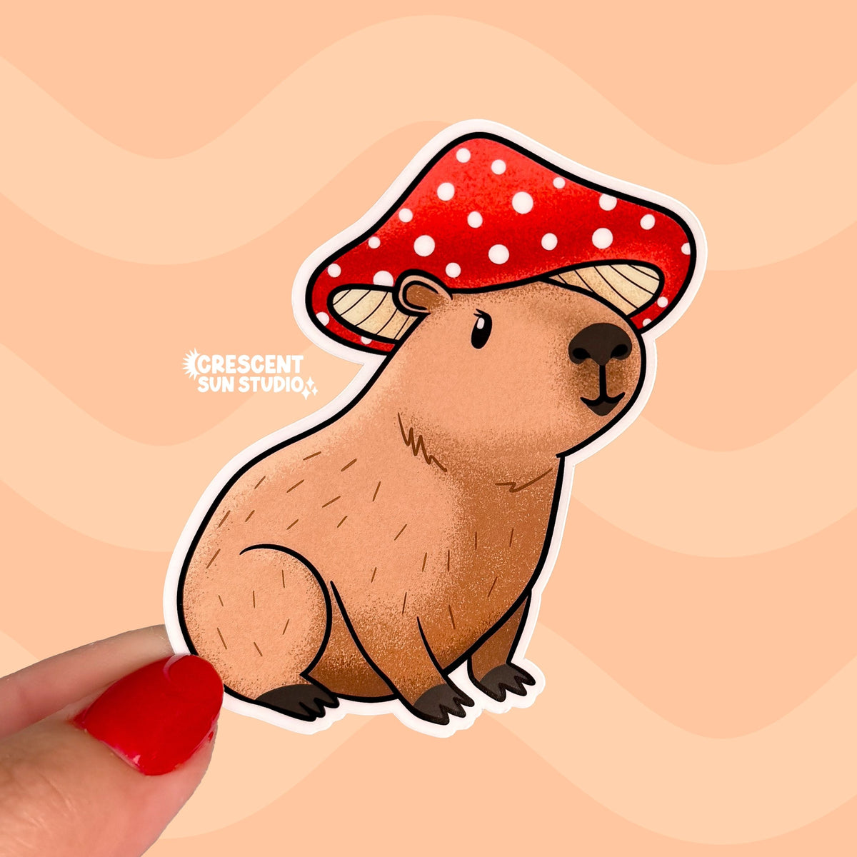 A sticker in the shape of a sitting capybara wearing a mushroom cap on his head. The capybara is brown and has a happy expression on his face. His cap is red with white spots.