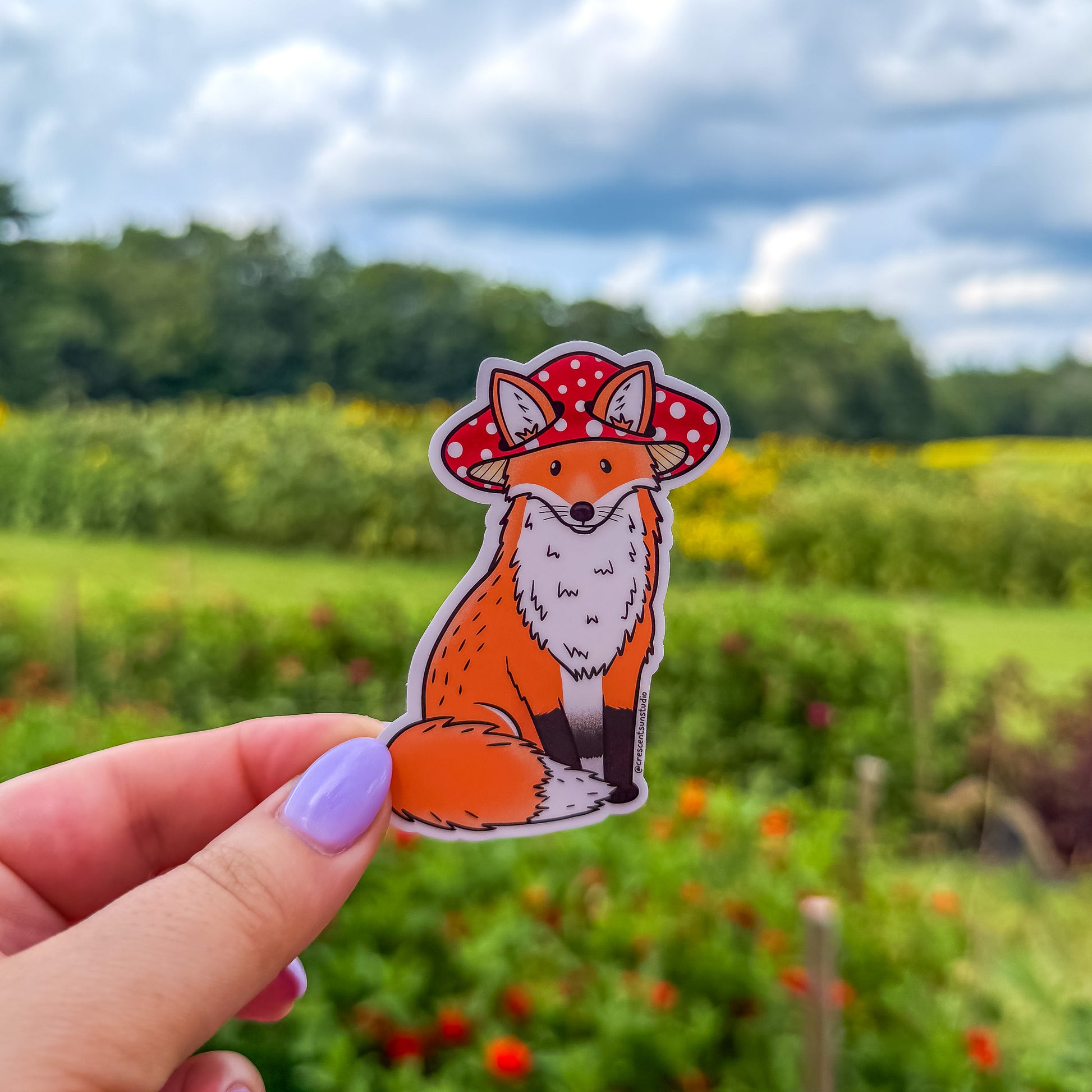 A die cut sticker of an orange fox wearing a red mushroom cap. The mushroom hat has holes for the fox's ears to peek through.
