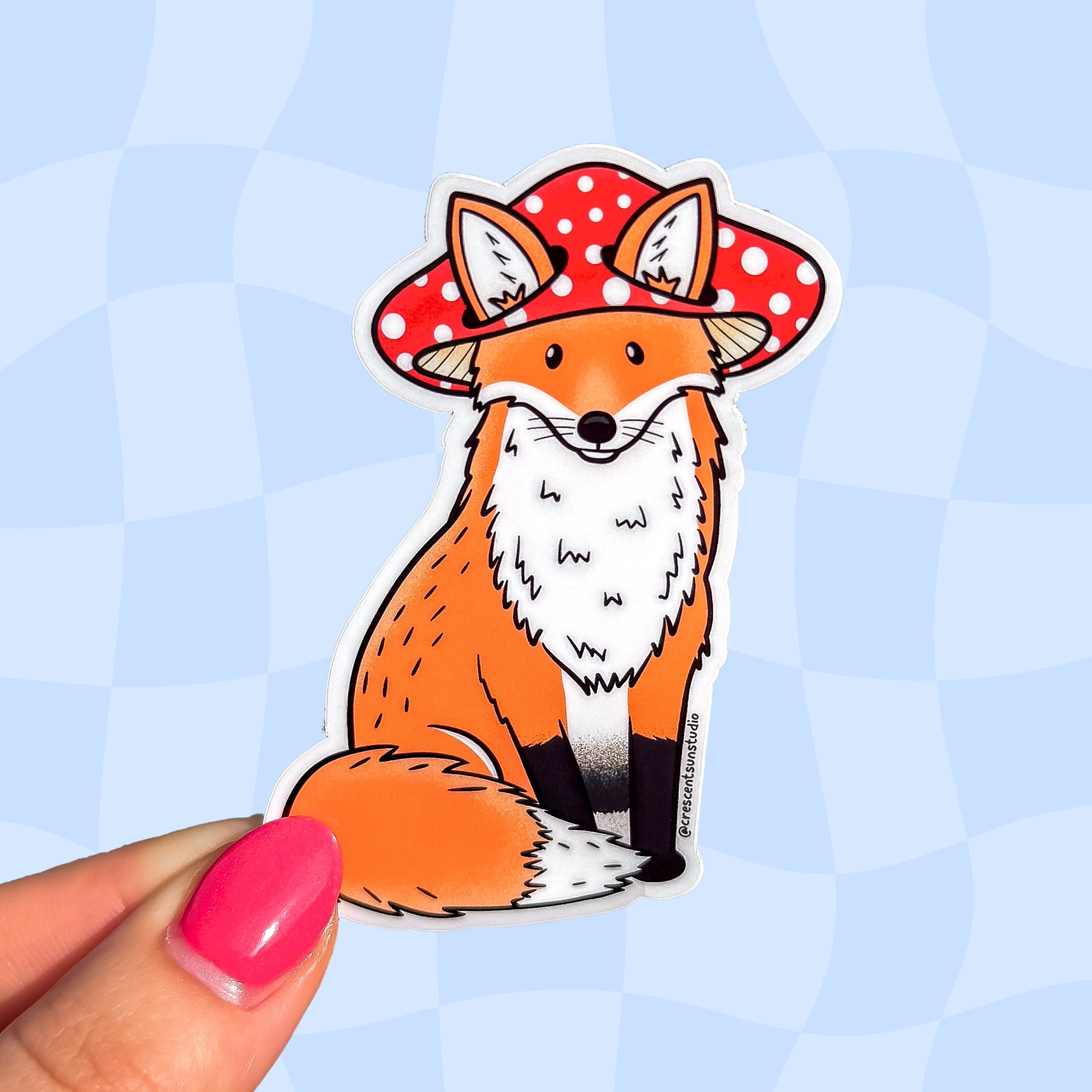 A die cut sticker of an orange fox wearing a red mushroom cap. The mushroom hat has holes for the fox's ears to peek through.