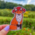 A die cut sticker of an orange fox wearing a red mushroom cap. The mushroom hat has holes for the fox's ears to peek through.