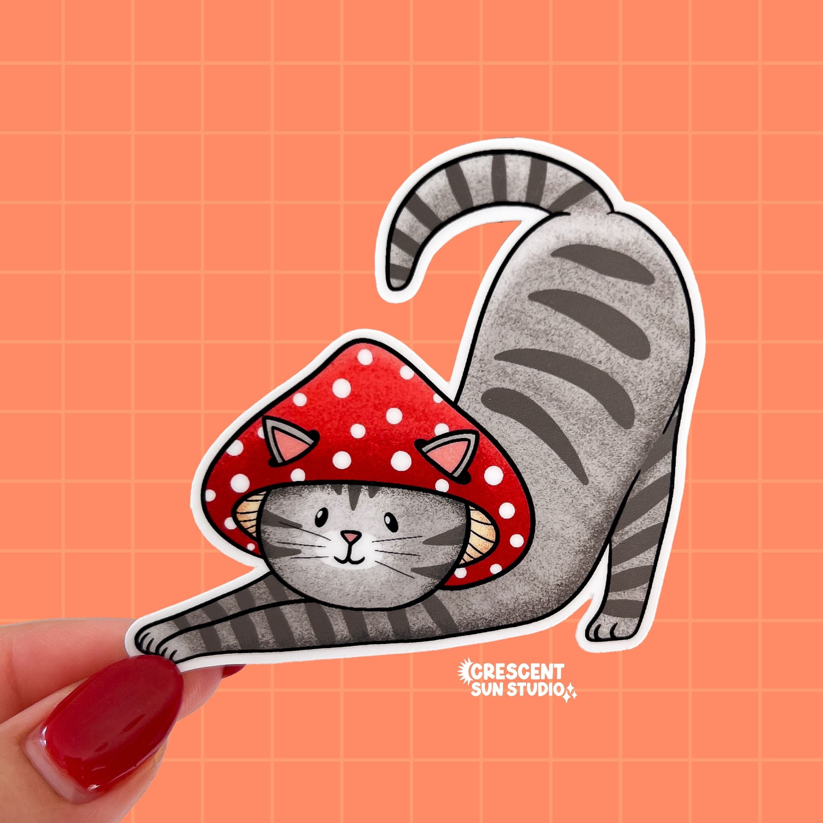 A sticker in the shape of a cat wearing a mushroom hat is held close to the camera. The cat is a grey tabby (medium grey with darker grey stripes) and has a sweet expression on his face. His red mushroom hat with white spots has spots for his ears to poke through. The cat is in a stretching pose with his tail curling up behind him.

In this image, a person holds the cat-shaped sticker up close to the camera.