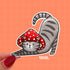 A sticker in the shape of a cat wearing a mushroom hat is held close to the camera. The cat is a grey tabby (medium grey with darker grey stripes) and has a sweet expression on his face. His red mushroom hat with white spots has spots for his ears to poke through. The cat is in a stretching pose with his tail curling up behind him.

In this image, a person holds the cat-shaped sticker up close to the camera.