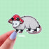 A die cut sticker of an opossum wearing a red and white mushroom cap for a hat.
