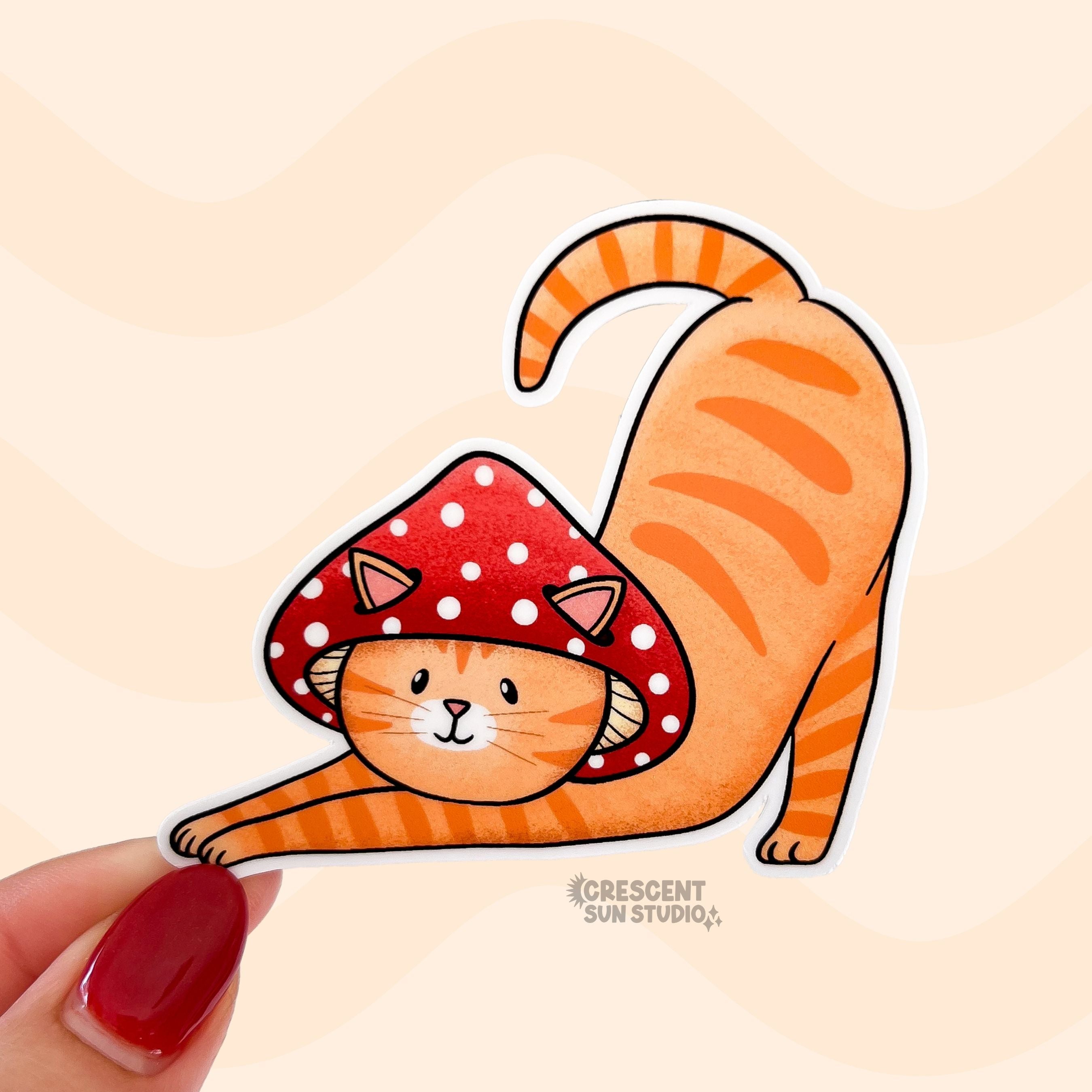 A sticker in the shape of a cat wearing a mushroom hat is held close to the camera. The cat is a tabby (orange with darker orange stripes) and has a sweet expression on his face. His red mushroom hat with white spots has spots for his ears to poke through. The cat is in a stretching pose with his tail curling up behind him.