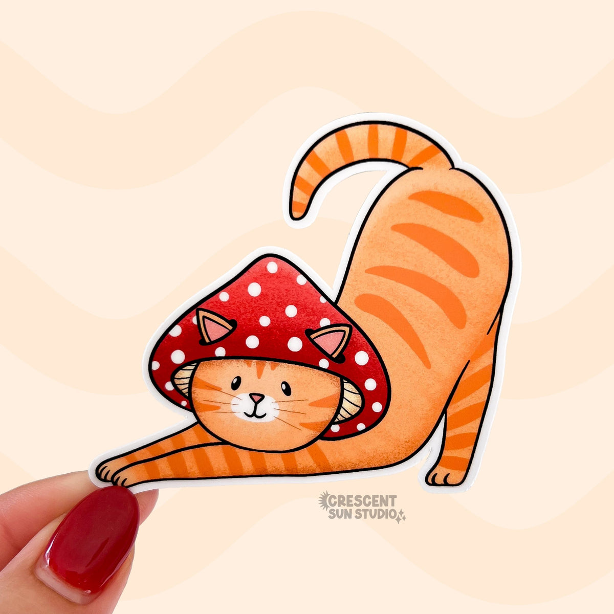 A sticker in the shape of a cat wearing a mushroom hat is held close to the camera. The cat is a tabby (orange with darker orange stripes) and has a sweet expression on his face. His red mushroom hat with white spots has spots for his ears to poke through. The cat is in a stretching pose with his tail curling up behind him.