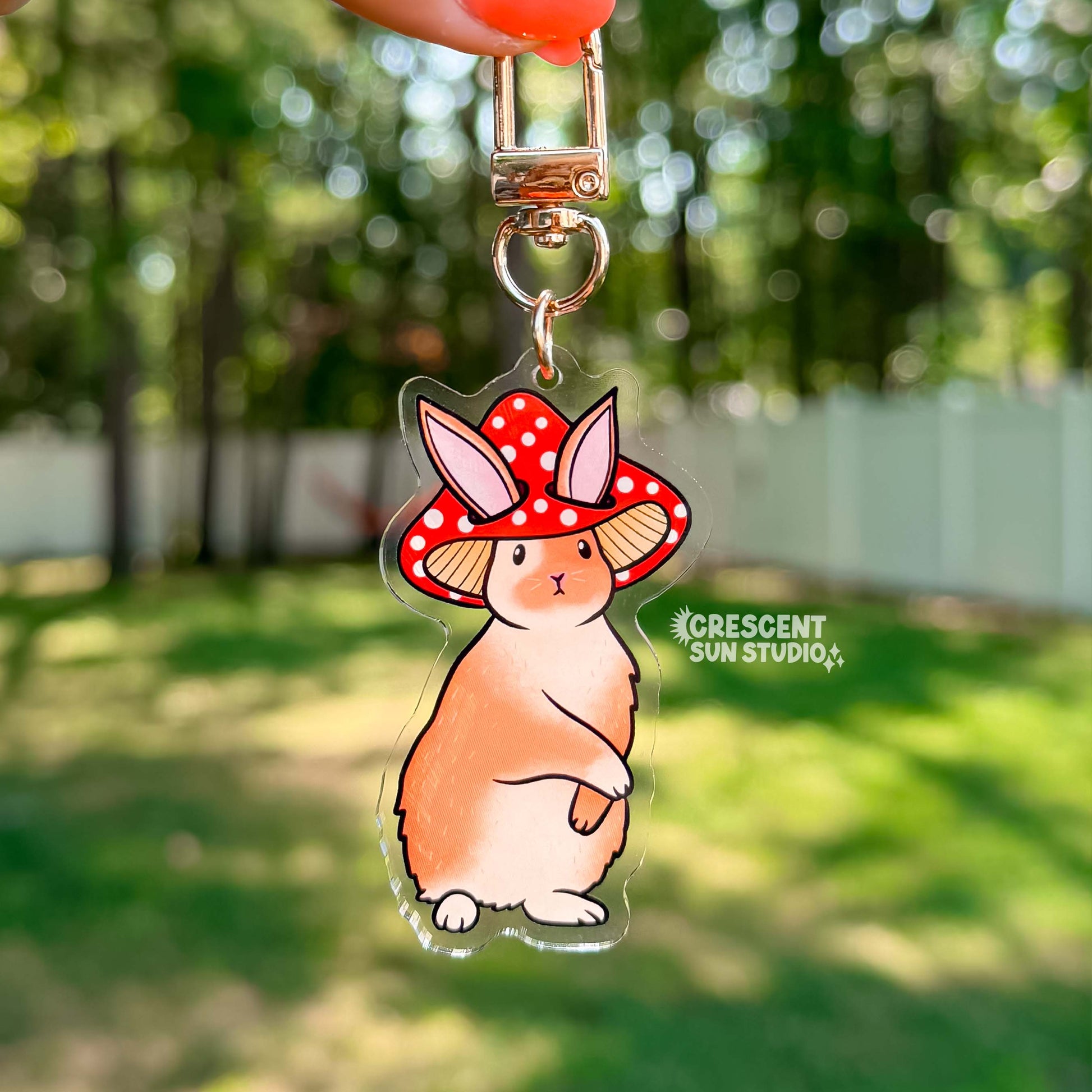 Acrylic die cut of a fluffy brown and white rabbit wearing a red spotted mushroom cap, standing upright with a composed expression. Keychain has a gold u-shaped clasp.