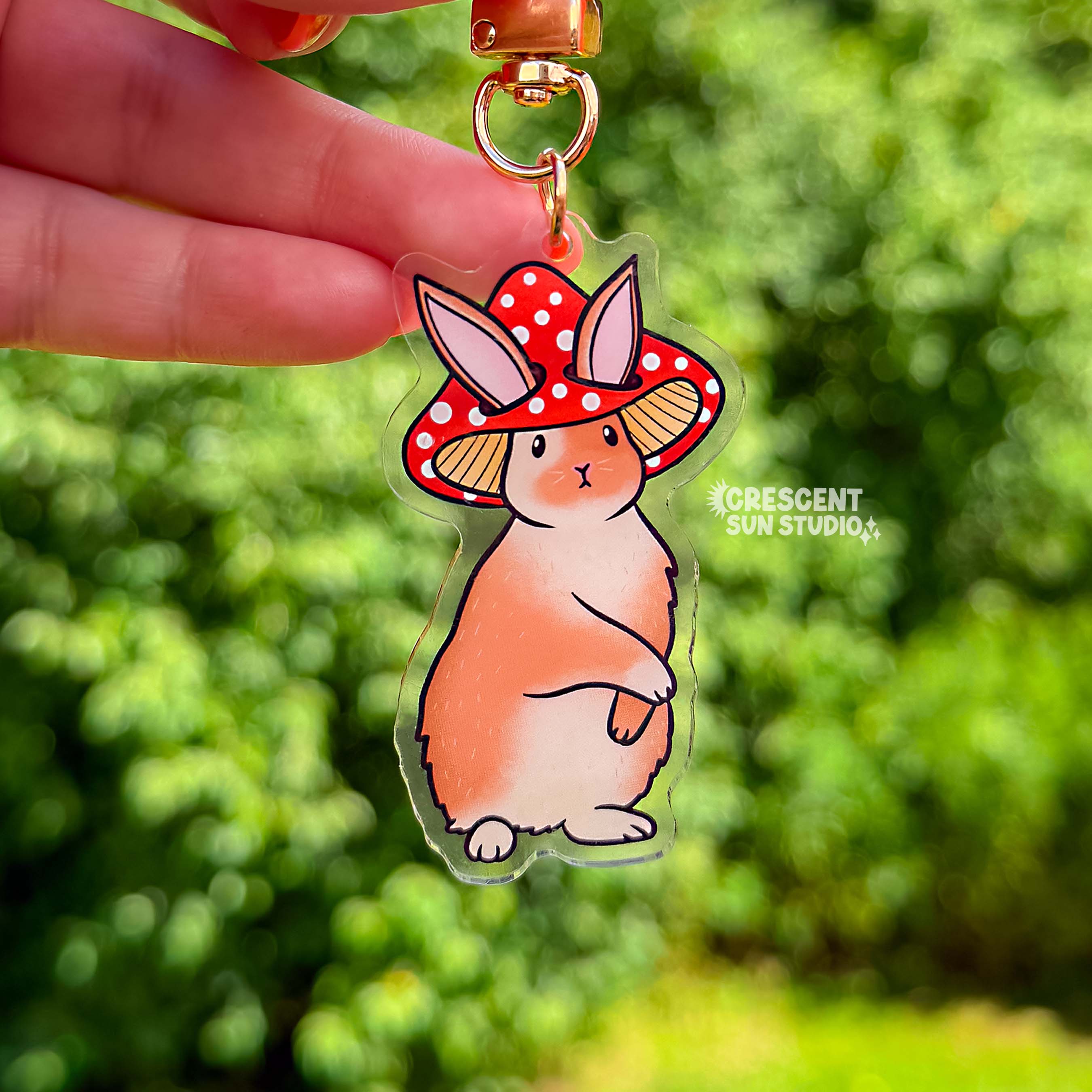 Acrylic die cut of a fluffy brown and white rabbit wearing a red spotted mushroom cap, standing upright with a composed expression. Keychain has a gold u-shaped clasp.