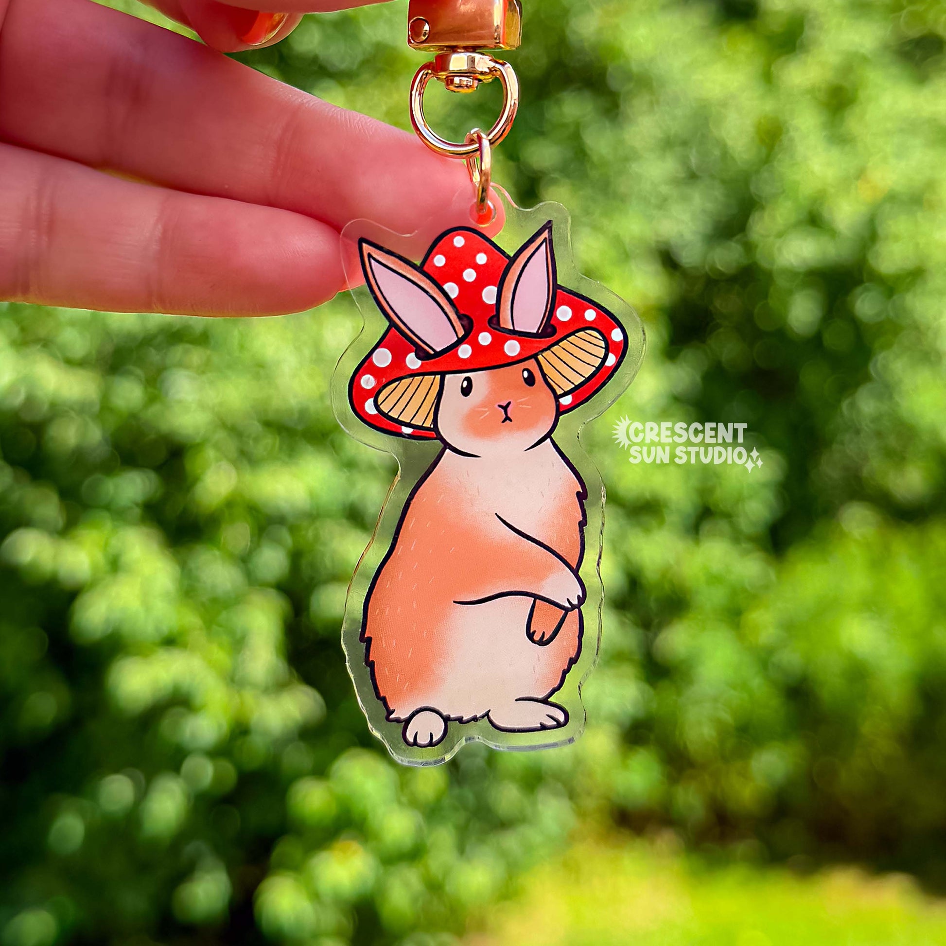 Acrylic die cut of a fluffy brown and white rabbit wearing a red spotted mushroom cap, standing upright with a composed expression. Keychain has a gold u-shaped clasp.