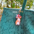 Acrylic die cut of a fluffy brown and white rabbit wearing a red spotted mushroom cap, standing upright with a composed expression. The keychain's gold u-shaped clasp is attached like a charm to the strap of a teal tote bag.