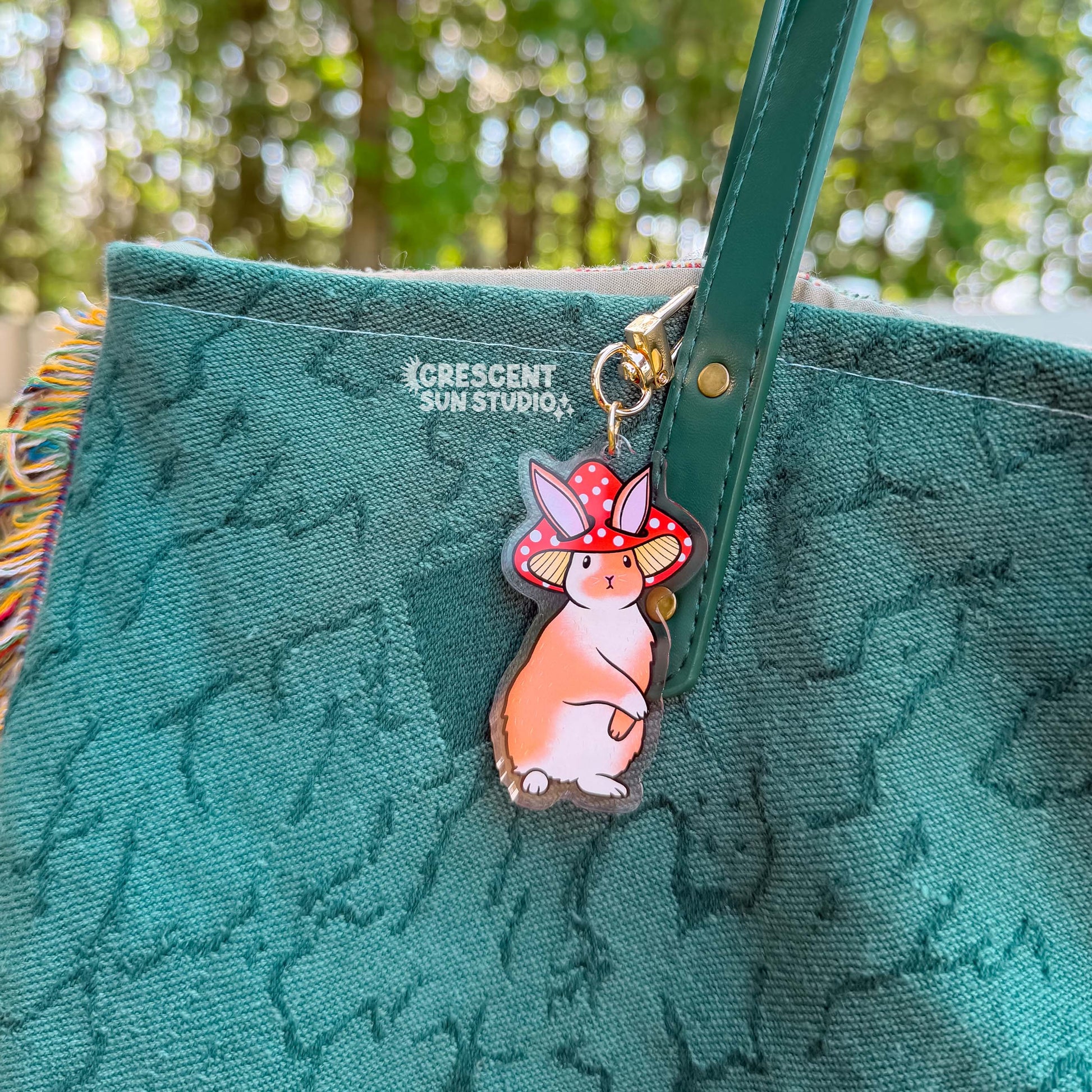 Acrylic die cut of a fluffy brown and white rabbit wearing a red spotted mushroom cap, standing upright with a composed expression. The keychain's gold u-shaped clasp is attached like a charm to the strap of a teal tote bag.