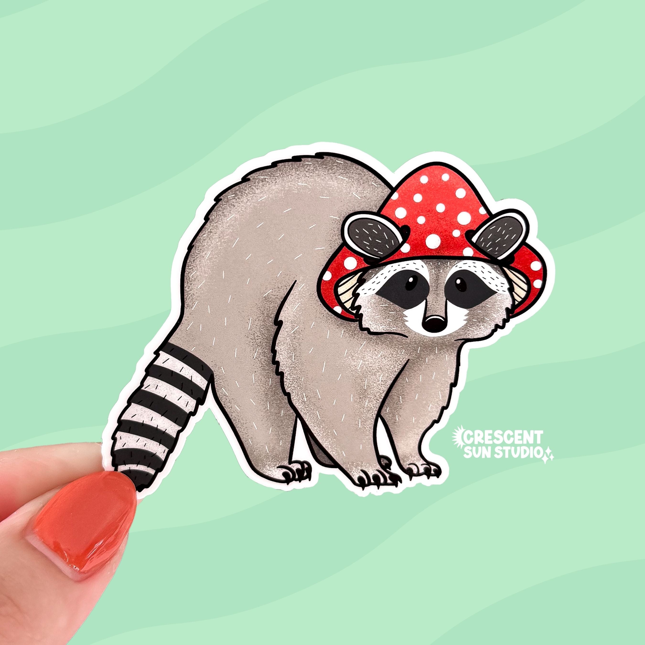 A diecut sticker of an adorable raccoon wearing a red mushroom cap.