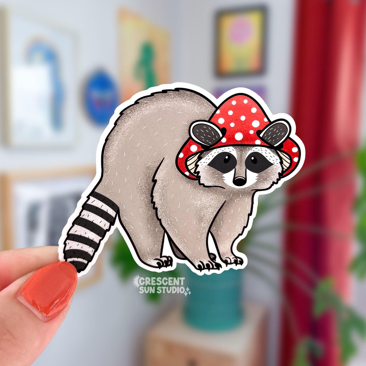 A diecut sticker of an adorable raccoon wearing a red mushroom cap.