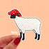 A die-cut sticker of a white wooly sheep with a black muzzle and legs. The sheep wears a red mushroom with white spots for a hat. The sheep's ears poke out through the hat.