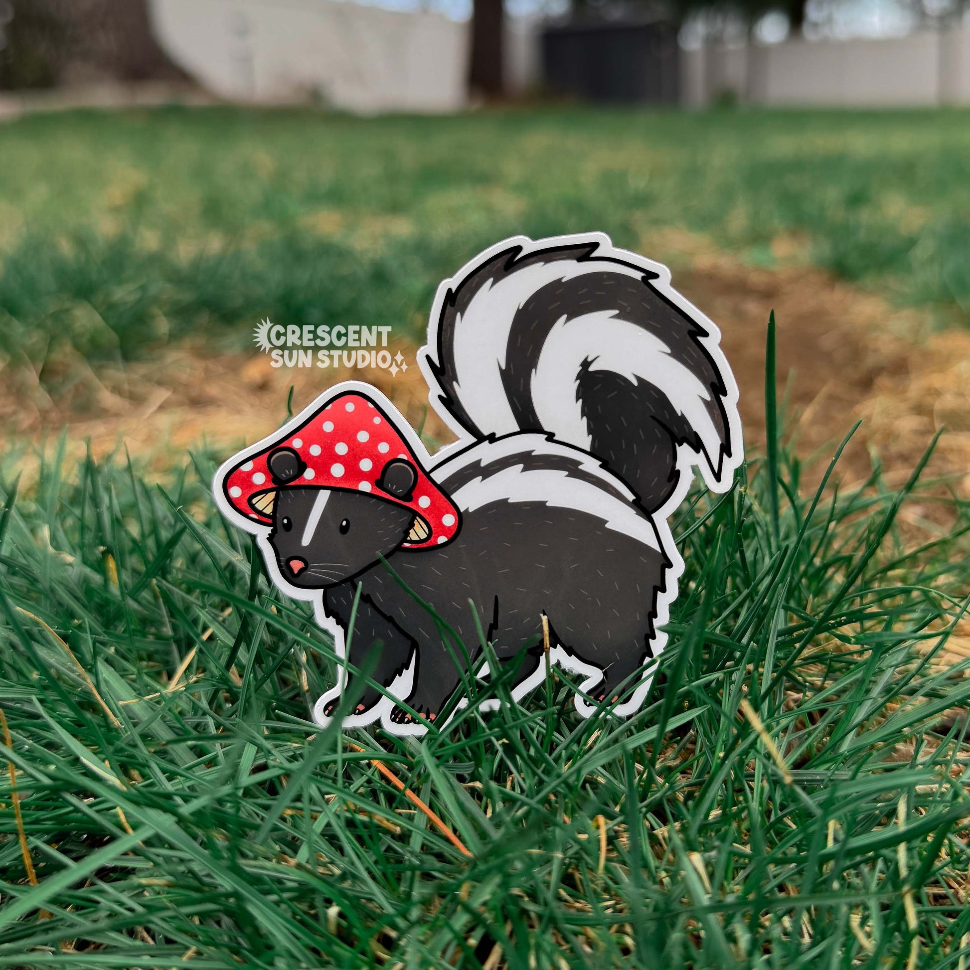 A die cut sticker of a skunk wearing a red and white mushroom cap for a hat. In this photo, the sticker is placed in a field of green grass.