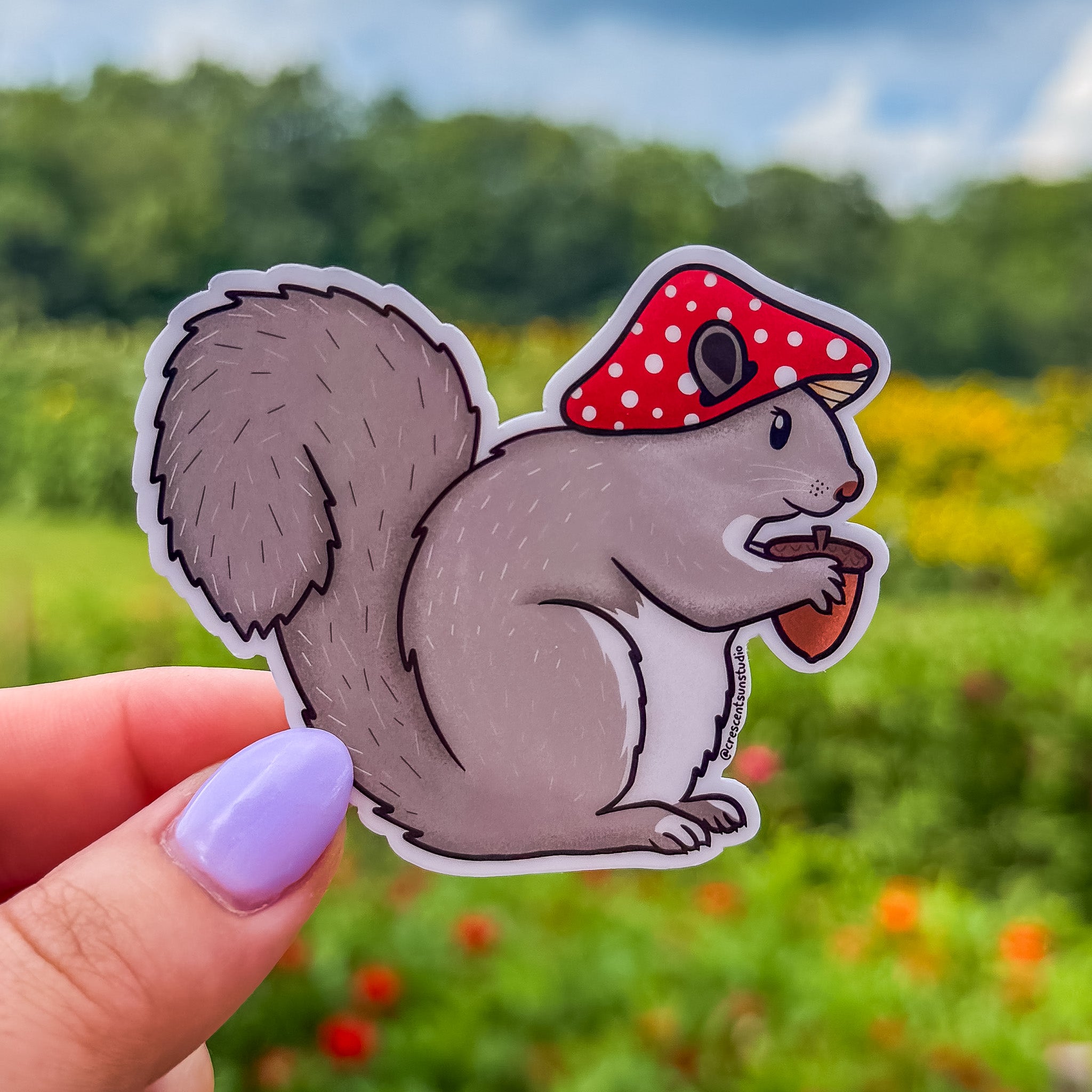 Die cut sticker of a squirrel wearing a red mushroom cap and holding an acorn
